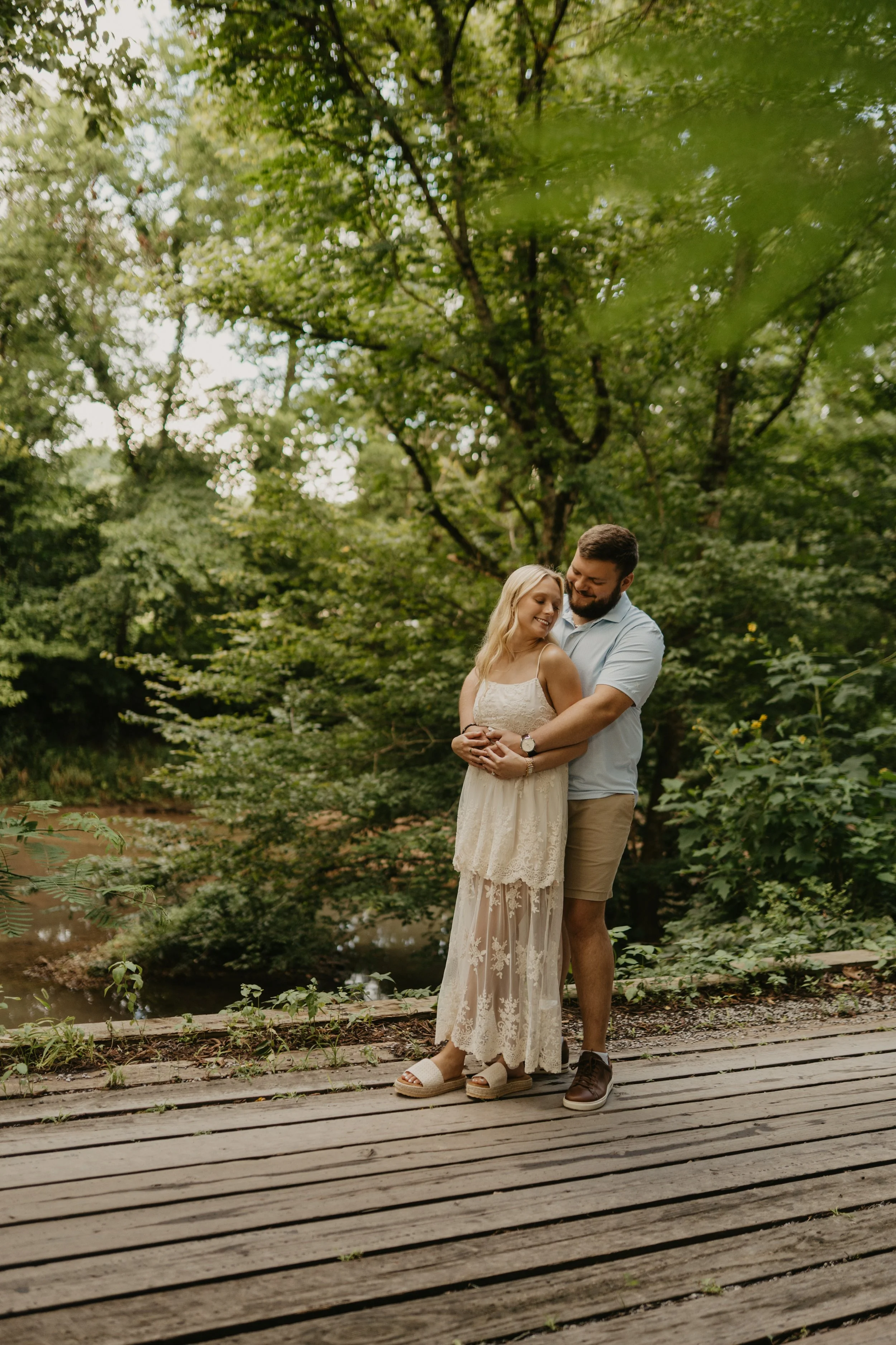 A couple standing on a wooden pathway in a lush green forest, embracing and smiling at each other.