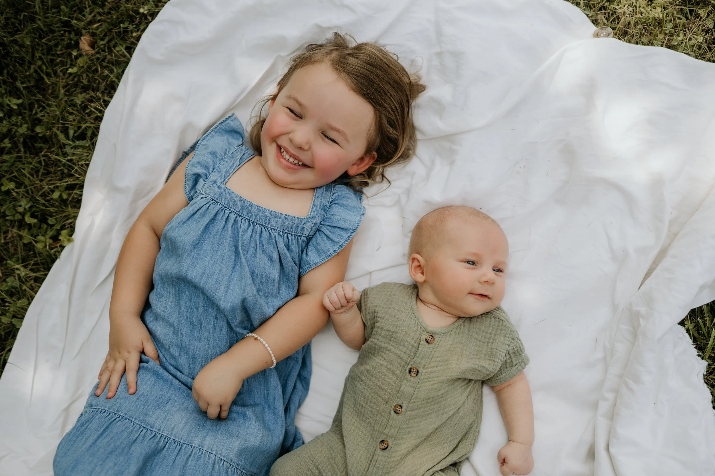 A young girl with curly hair and a blue dress lying on a white blanket outdoors, smiling with eyes closed, next to a baby in a green shirt also lying on the blanket.