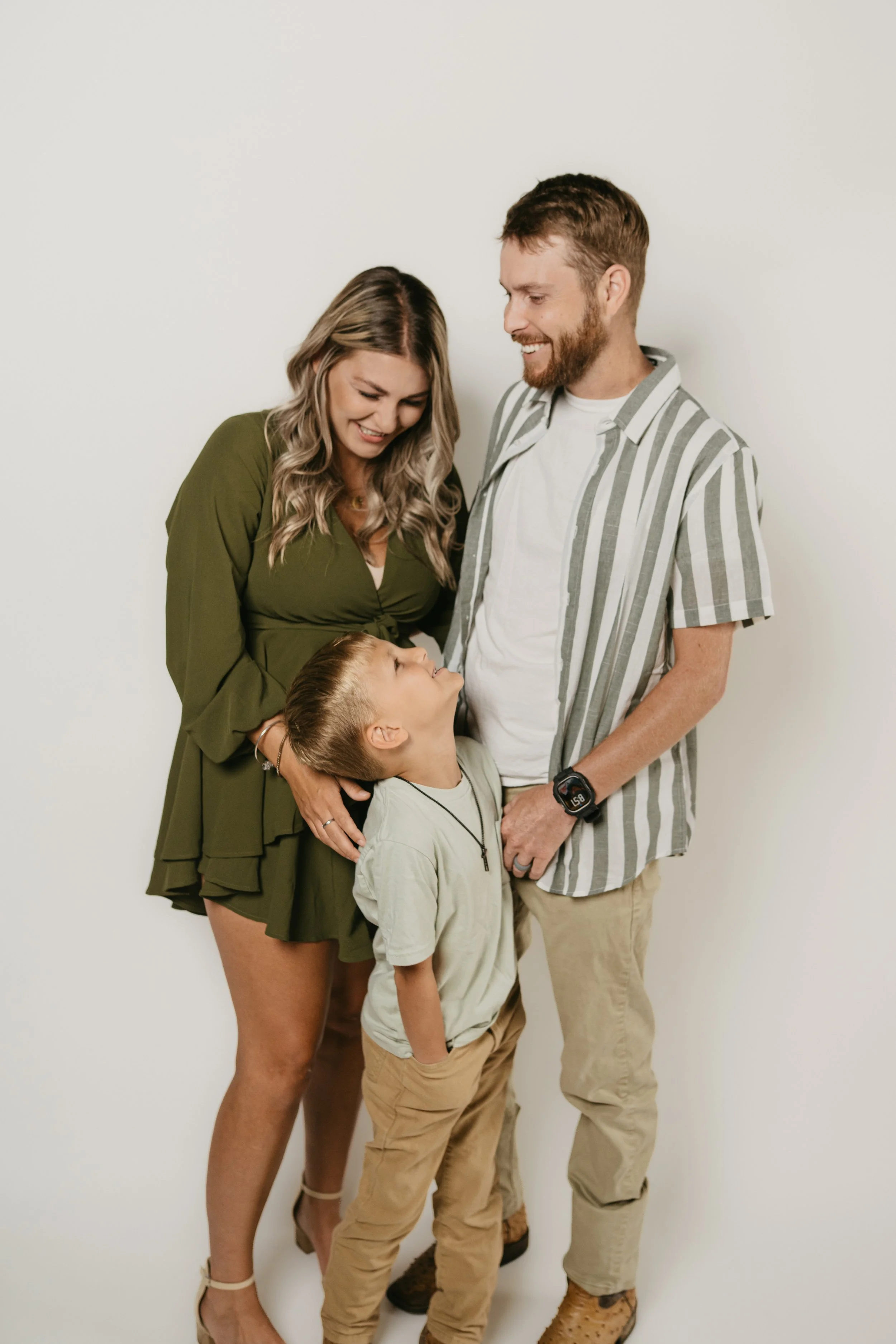 Family of four smiling and looking at each other against a plain white wall. The mother has long wavy hair and is wearing an olive green dress, the father has a beard and is wearing a striped shirt, and the two children, a boy and a girl, are looking