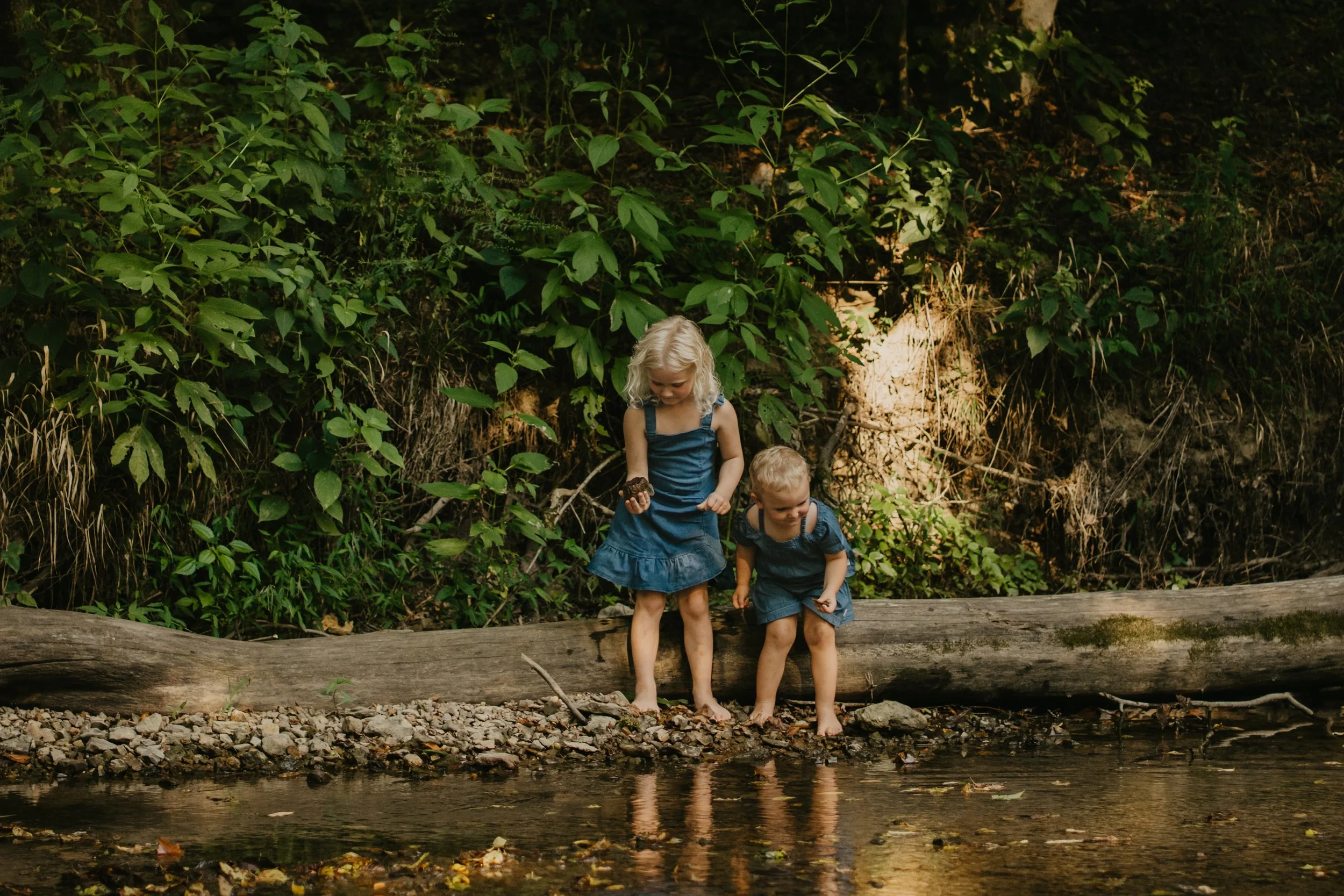 Two young children, a girl and a boy, standing on a log by a river in a lush, green wooded area. The girl is holding a rock, and both children are barefoot.