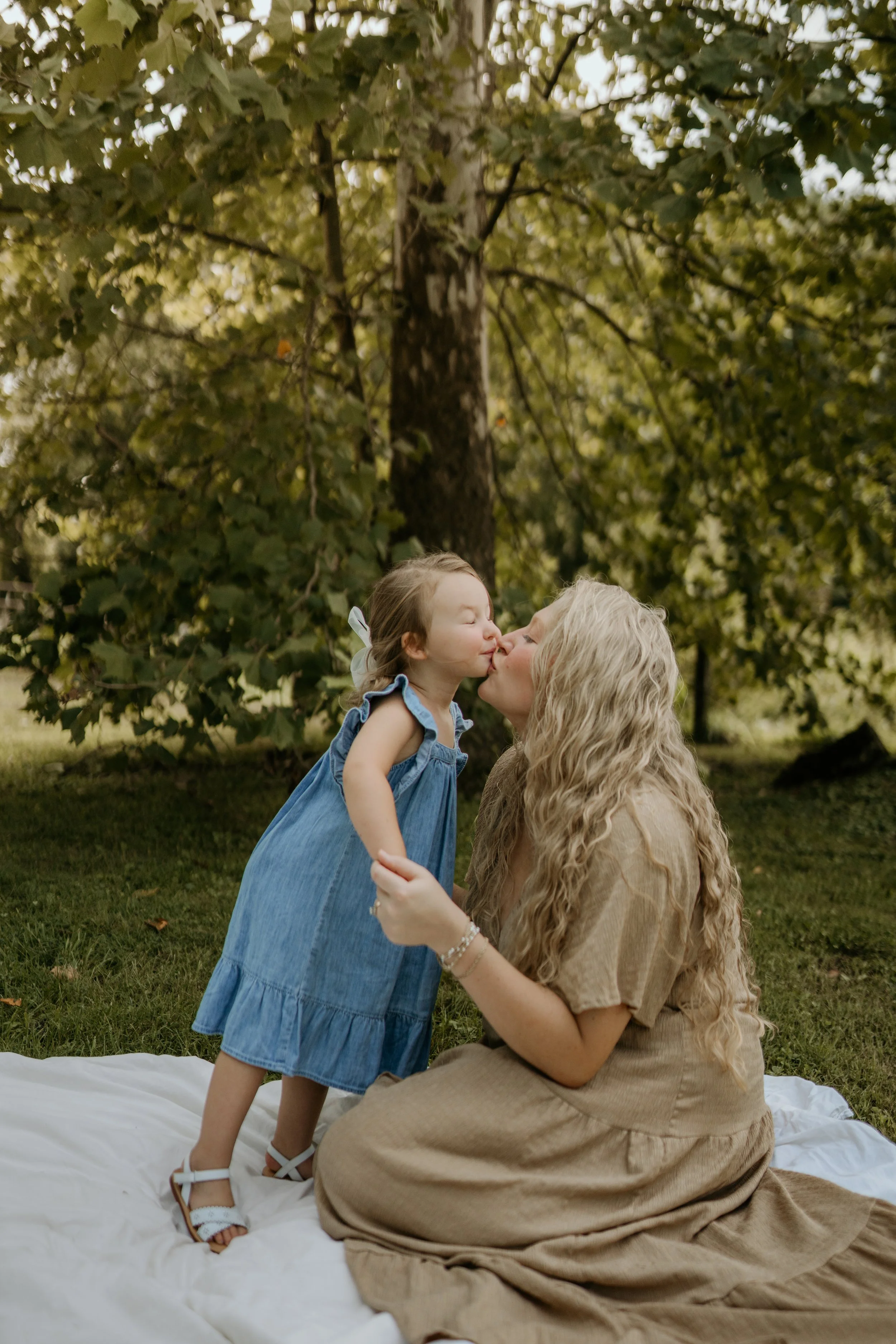 A woman with long, curly blonde hair and wearing a beige dress is kneeling on a white blanket on grass, holding a young girl by her waist. The girl, wearing a blue denim dress and white sandals, is standing and leaning in to kiss the woman on the lip