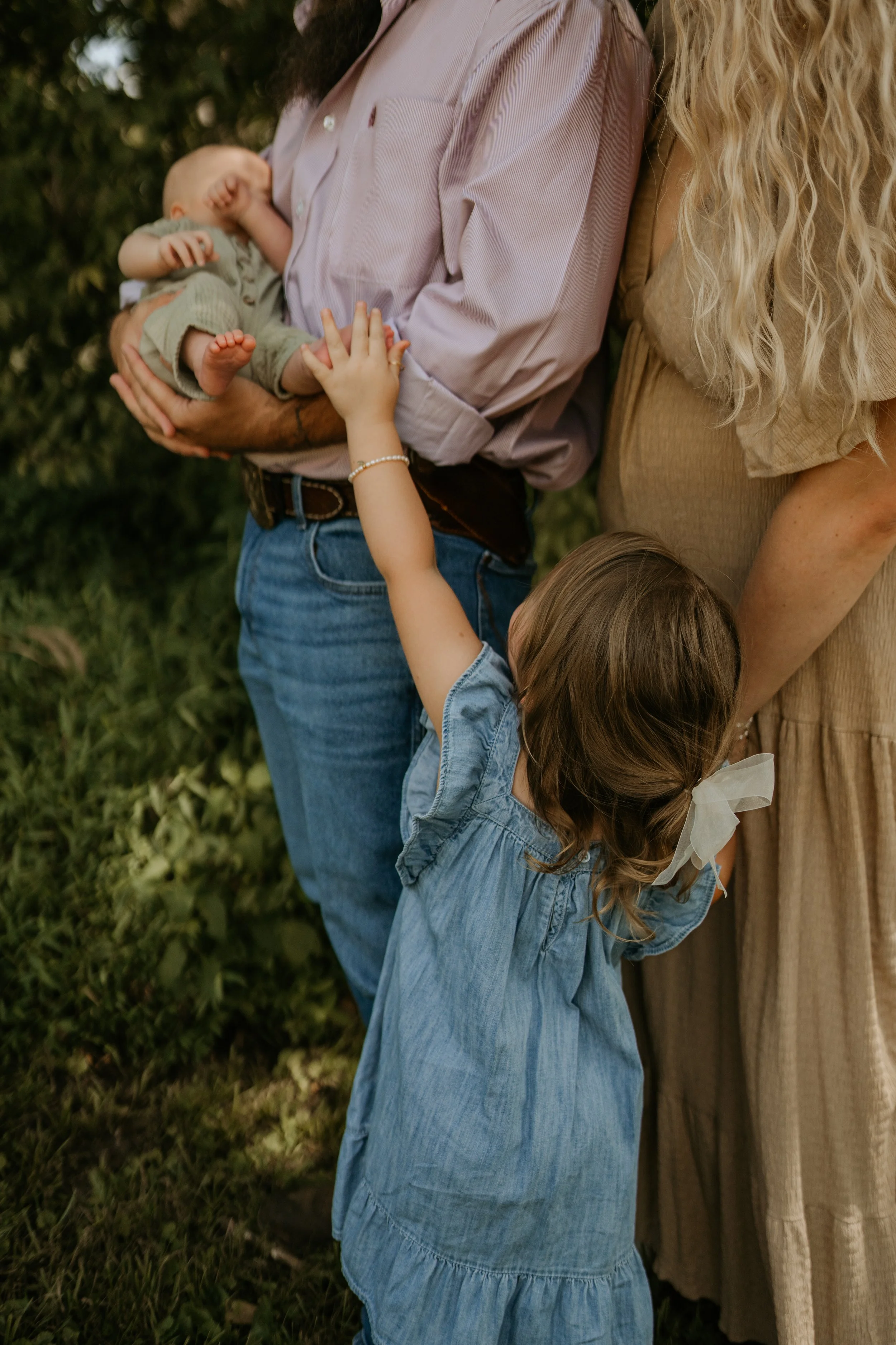 A man holding a sleeping baby while a woman and a young girl embrace in a grassy outdoor setting.