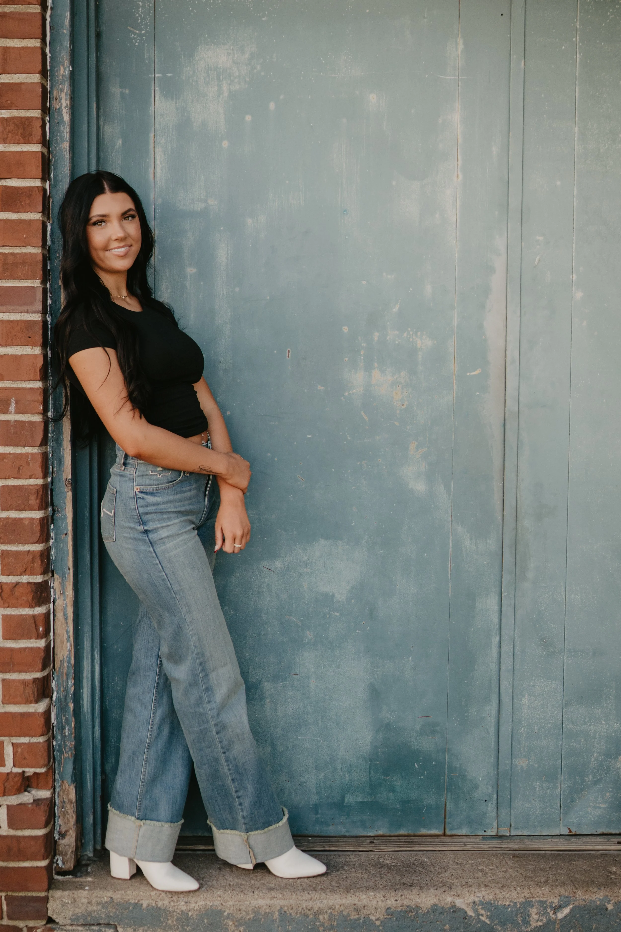 A woman with long black hair wearing a black crop top, high-waisted jeans, and white cowboy boots is standing against a teal metal door, with a brick wall on the left side of the image.