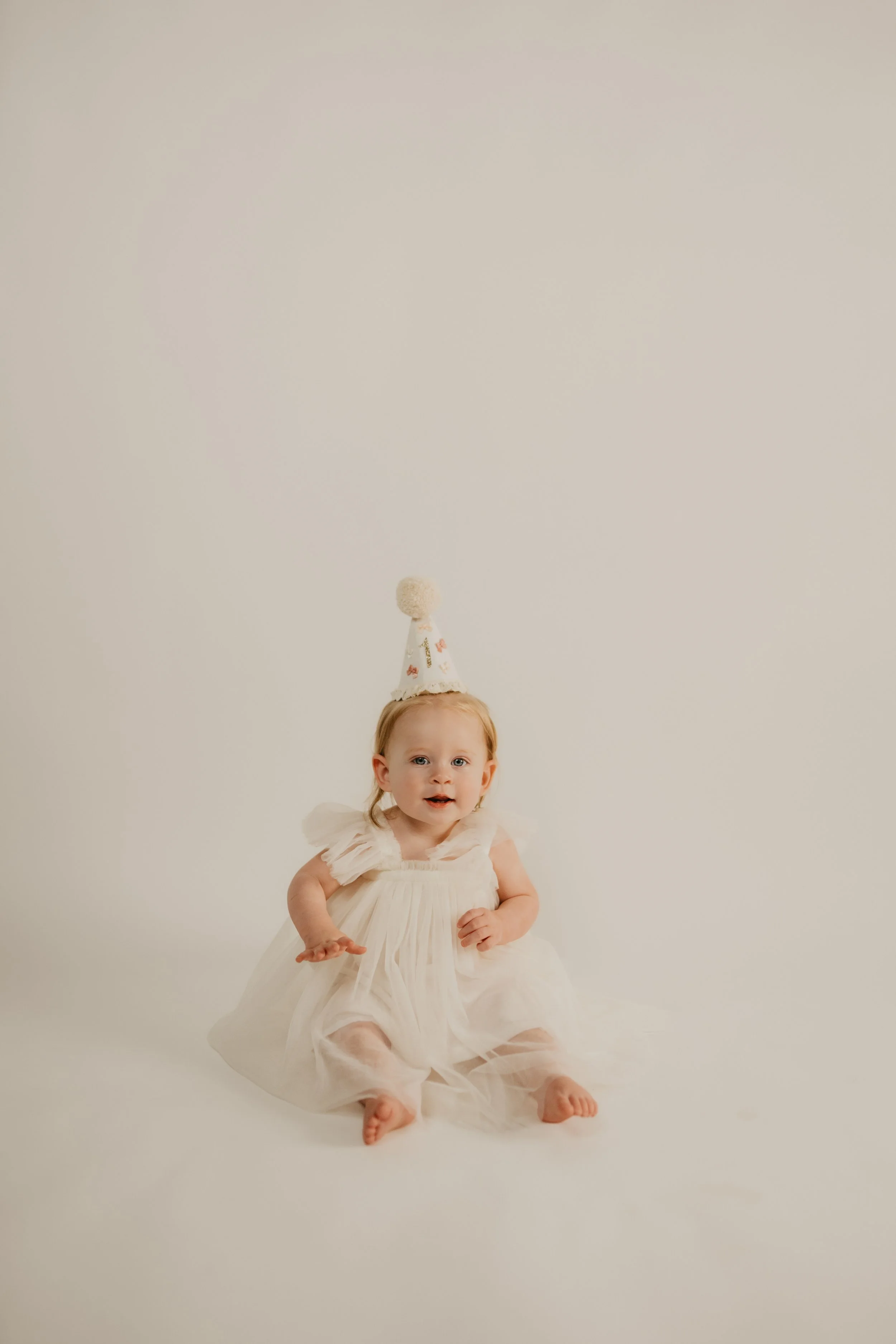 A young child in a white dress sitting on the floor, with a white background and a small party hat on their head.