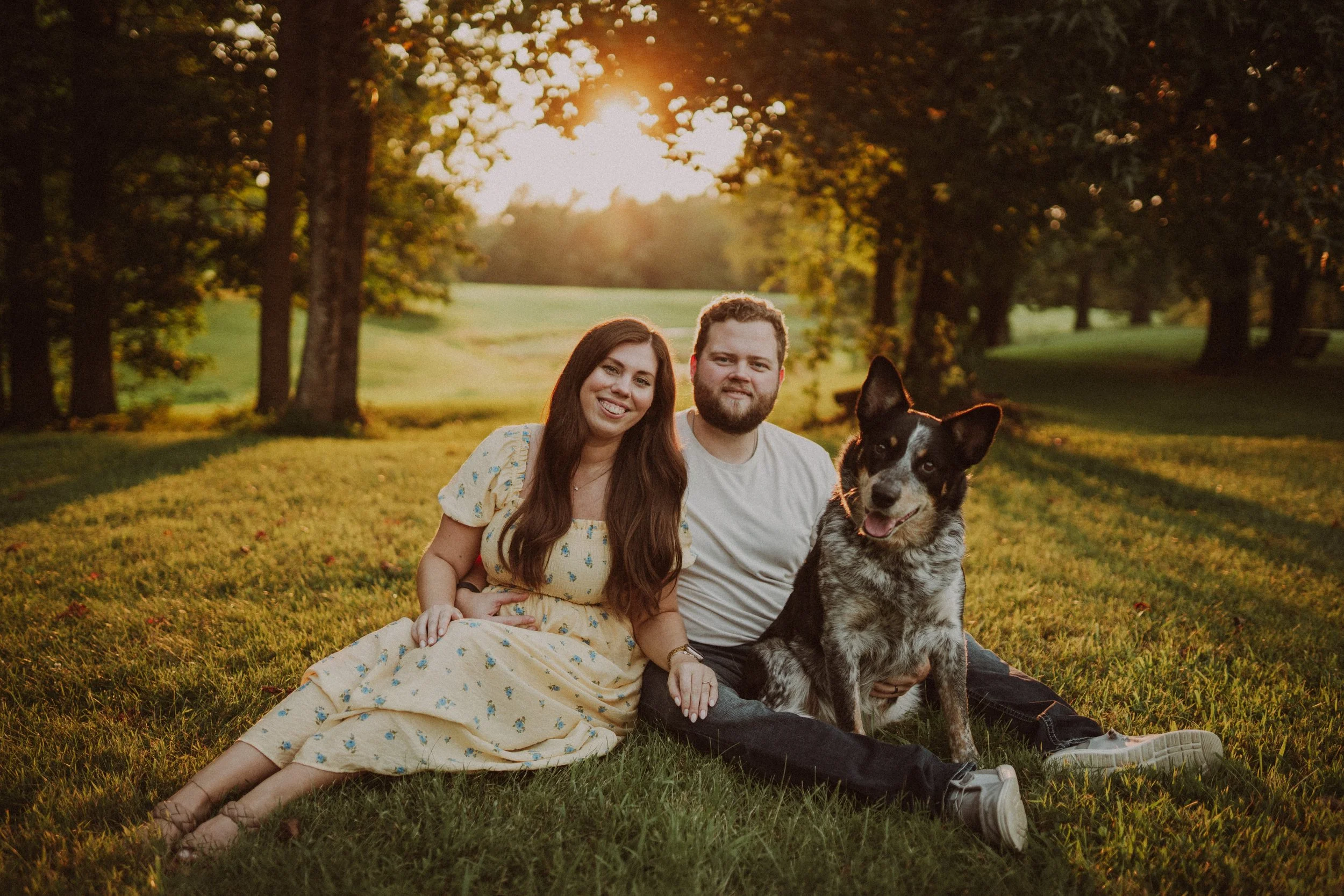 A young couple sitting on the grass in a park during sunset, with their dog, smiling and enjoying each other's company.