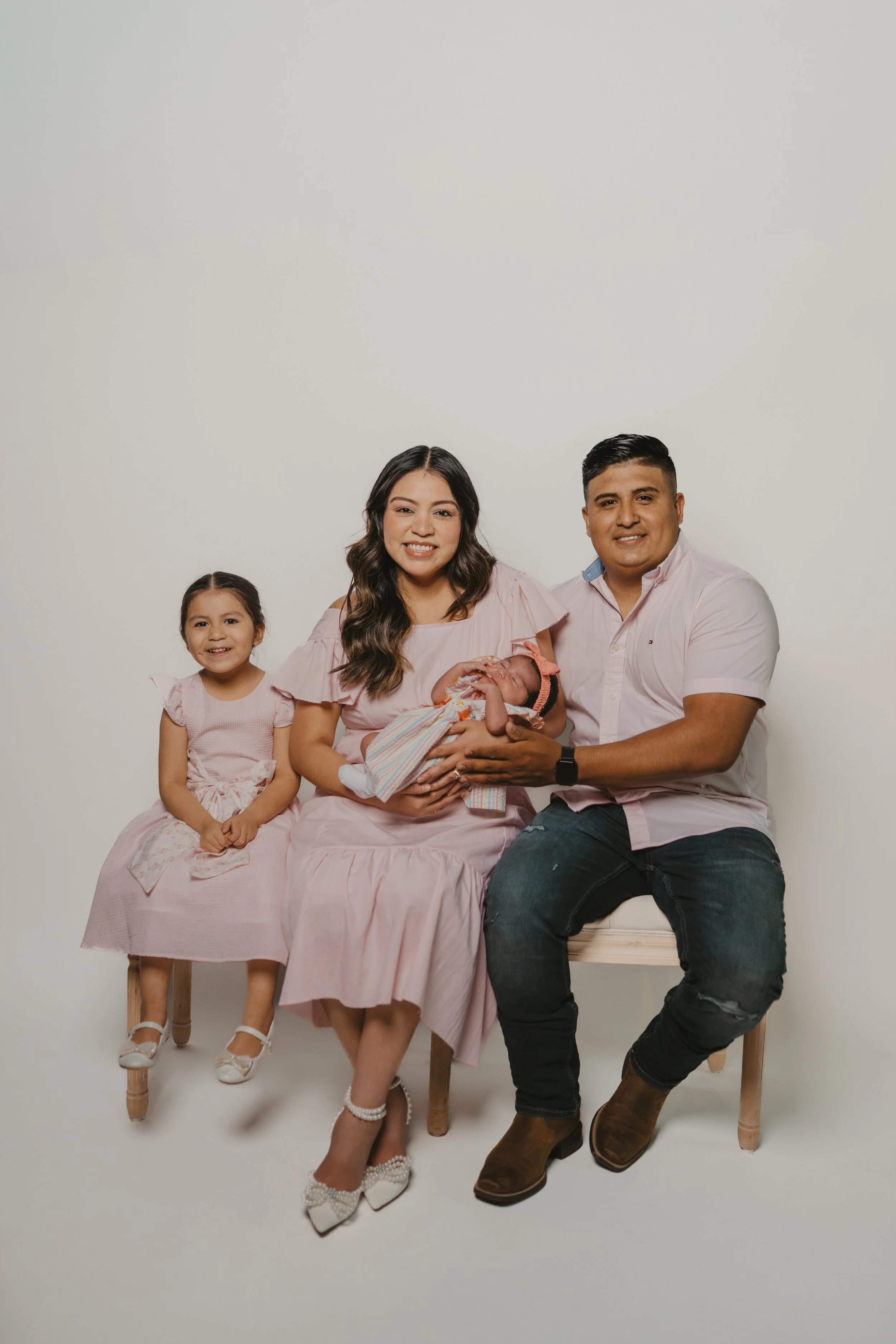 A smiling family of five, including a man, a woman, two daughters, and a newborn baby, sitting against a plain white background. The woman holds the baby wrapped in a blanket, and the family is dressed in pink and light colors.