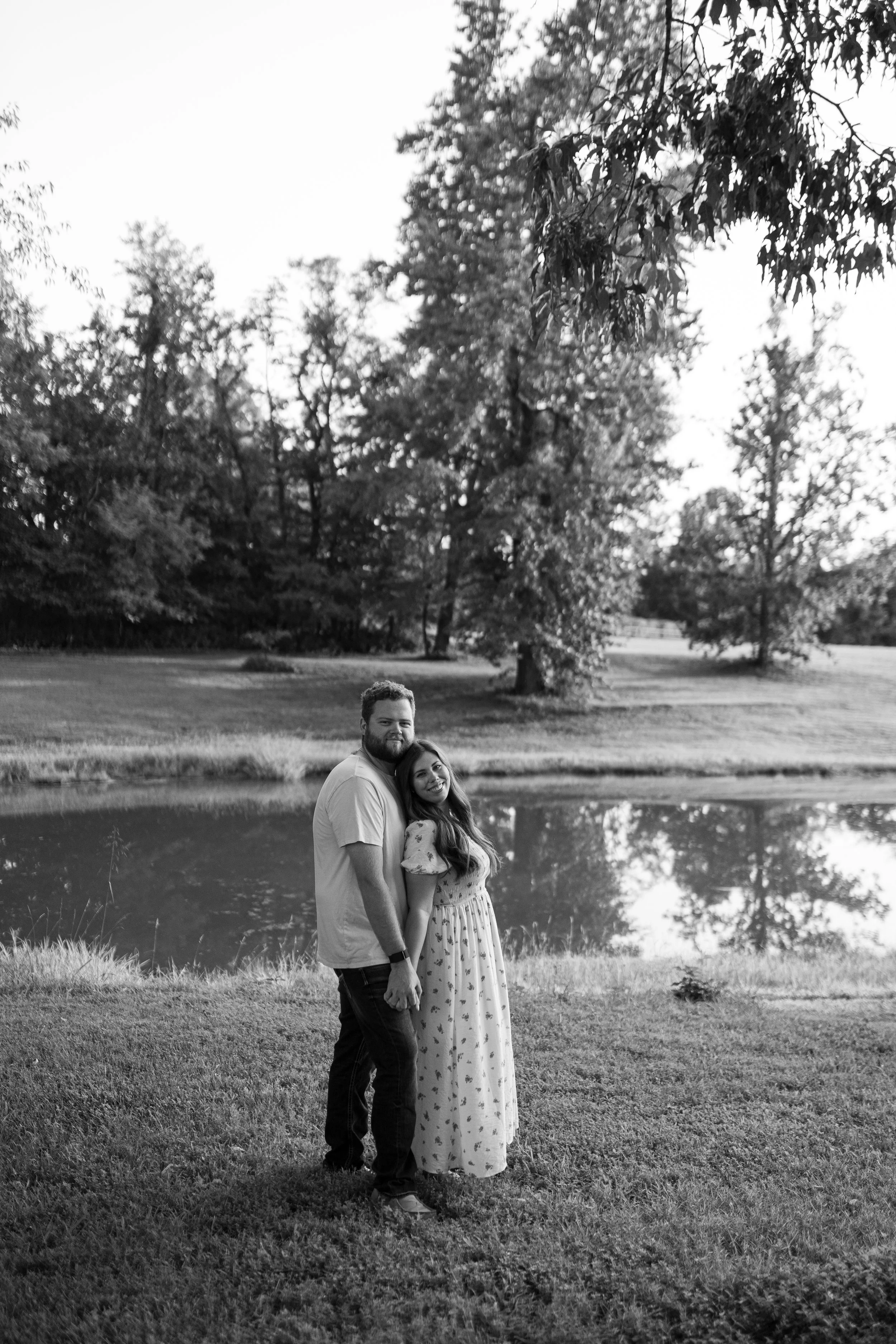 A black and white photo of a couple standing by a pond in a park with trees in the background. The man has a beard and is wearing a light-colored shirt; the woman is wearing a long, patterned dress with long hair. They are holding hands and smiling a
