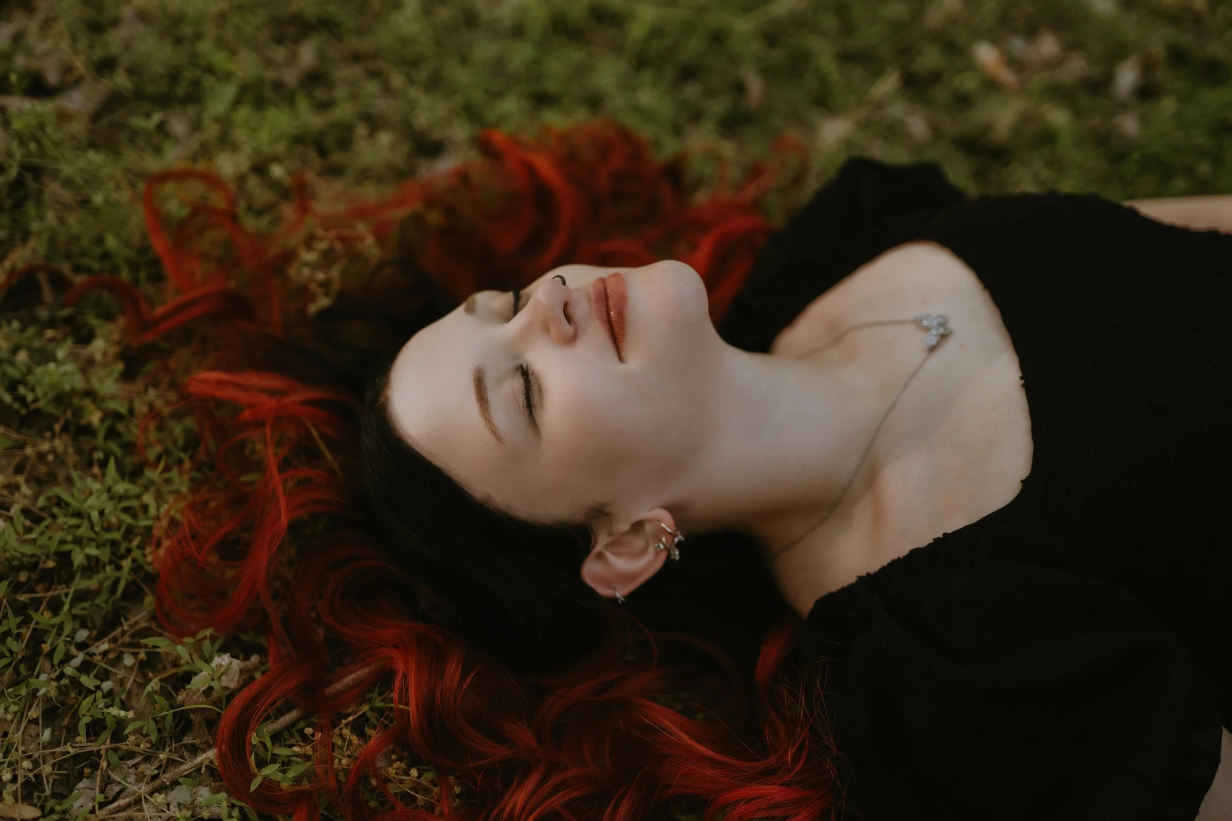 A woman with dark hair and red streaks lying on the grass with her eyes closed, wearing a black top and a necklace with a floral pendant.