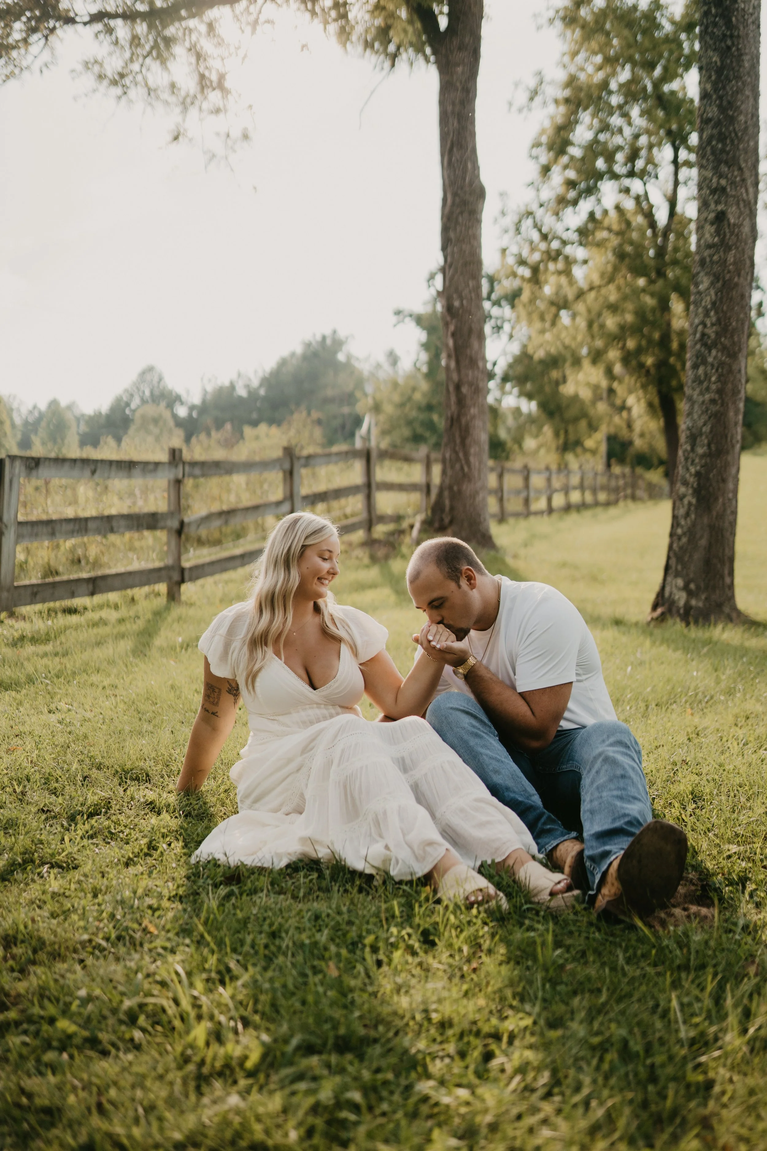 A man kneeling and kissing a woman's hand while sitting on the grass in a park, with trees and a wooden fence in the background.