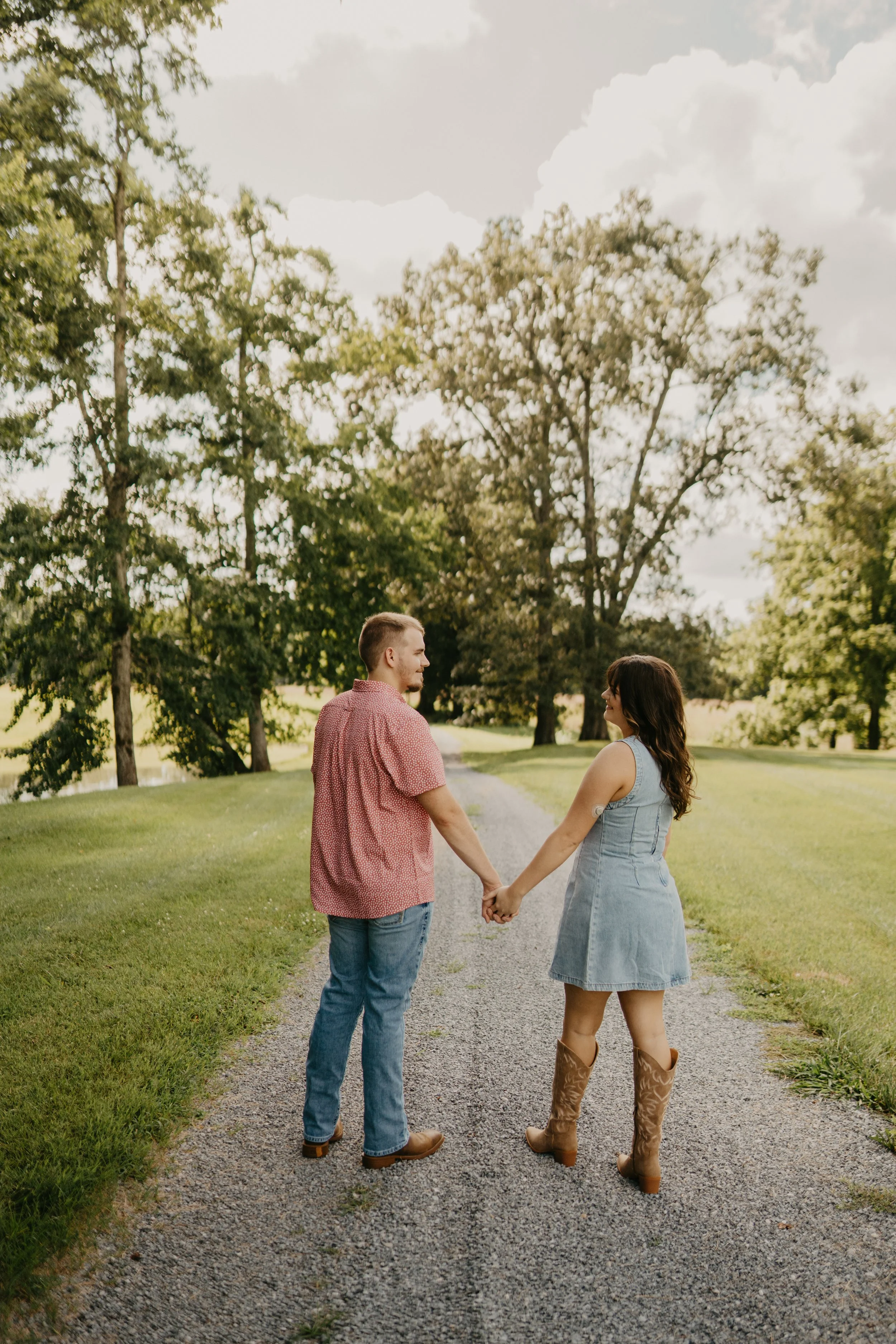 A couple holding hands and facing each other on a gravel path in a park-like setting, with trees and a cloudy sky in the background.