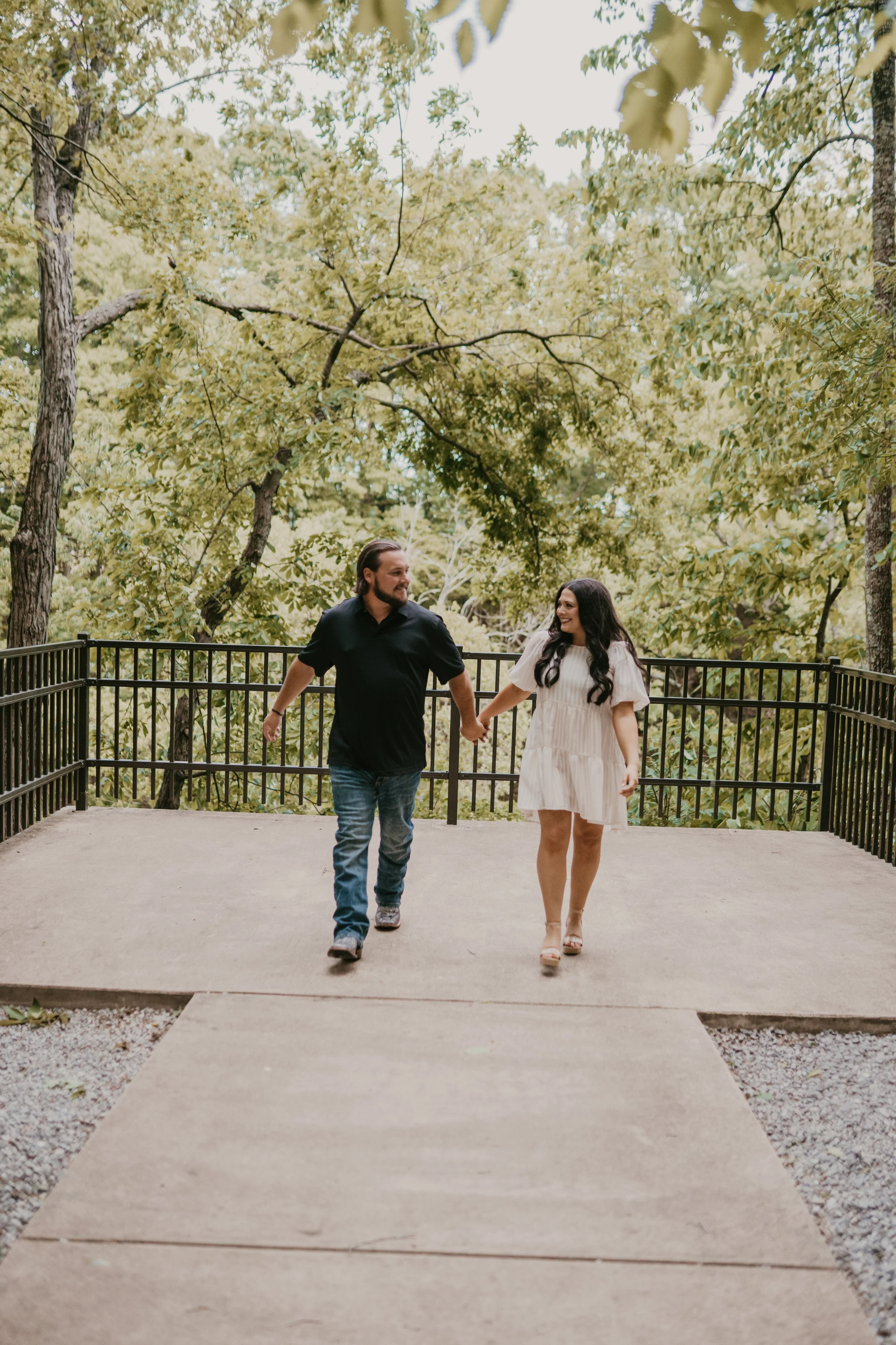 A man and woman holding hands while walking on a pathway in a park with green trees in the background.