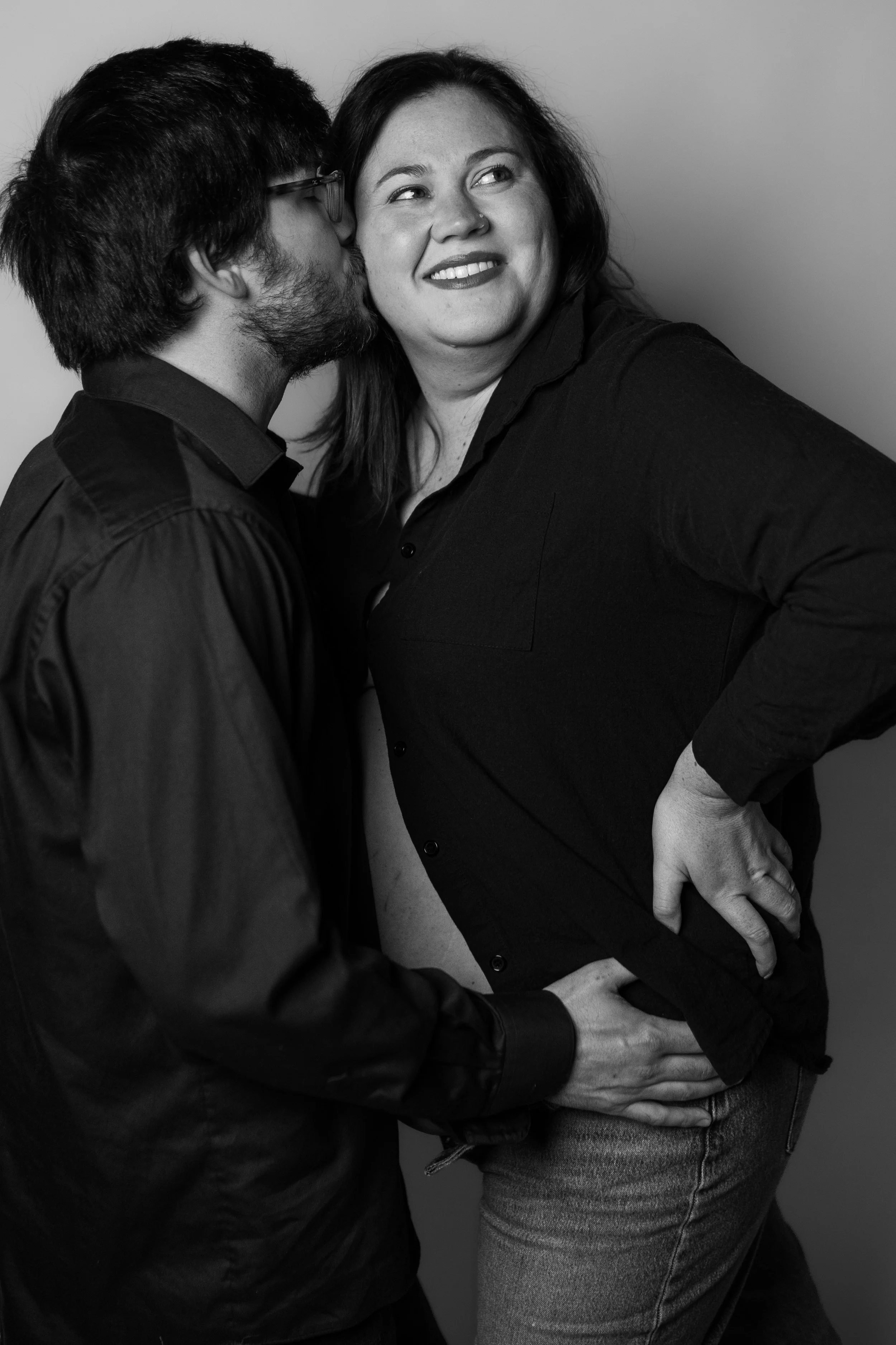 A man and a woman sharing an intimate moment, with the man whispering into the woman's ear as she smiles, wearing casual clothes, against a plain background.