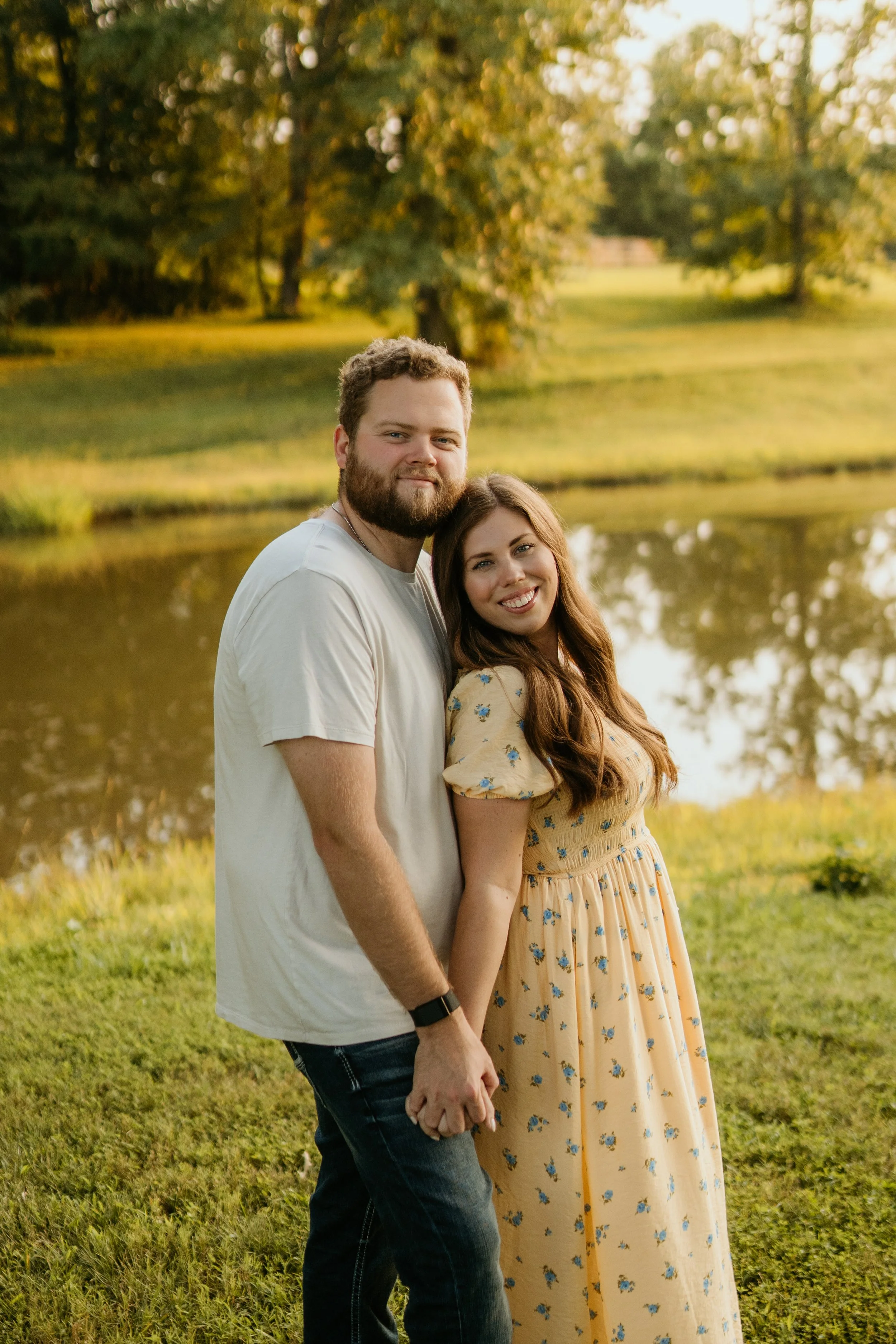 A couple holding hands by a lake during sunset, with trees in the background.