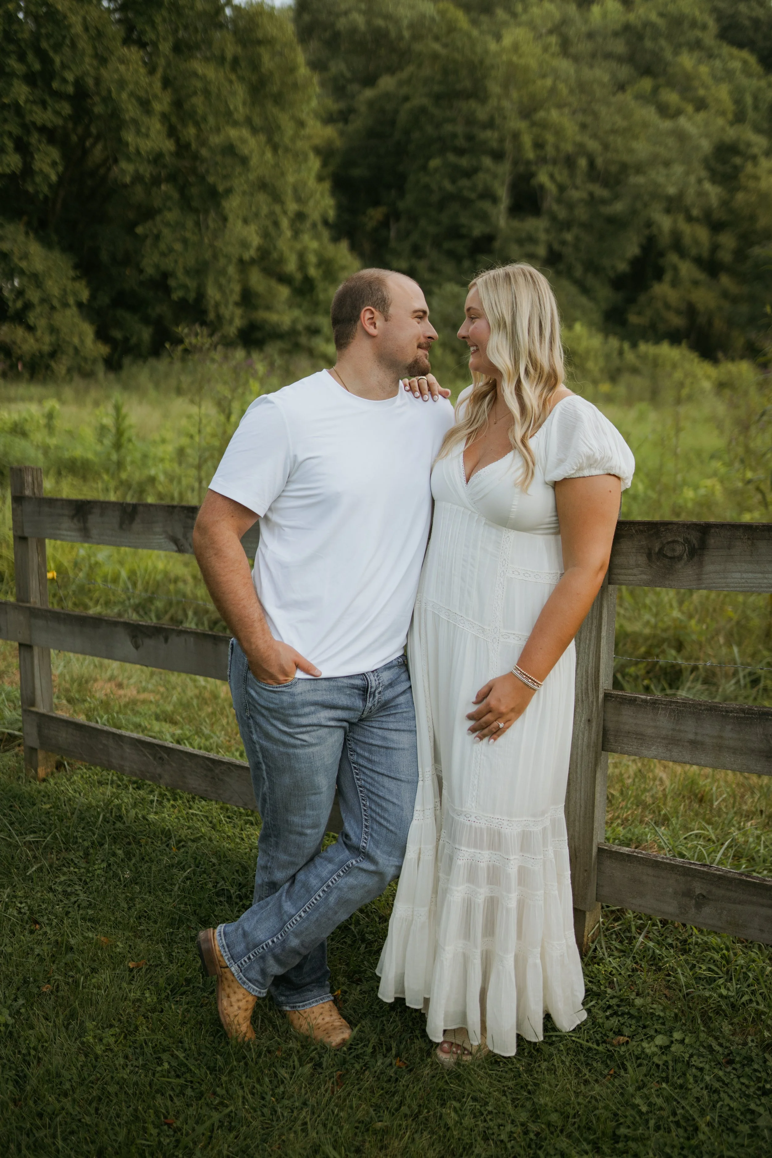 A couple standing outdoors by a wooden fence, smiling and looking at each other. The man is wearing a white t-shirt and jeans, and the woman is wearing a white dress. There are green trees and grass in the background.