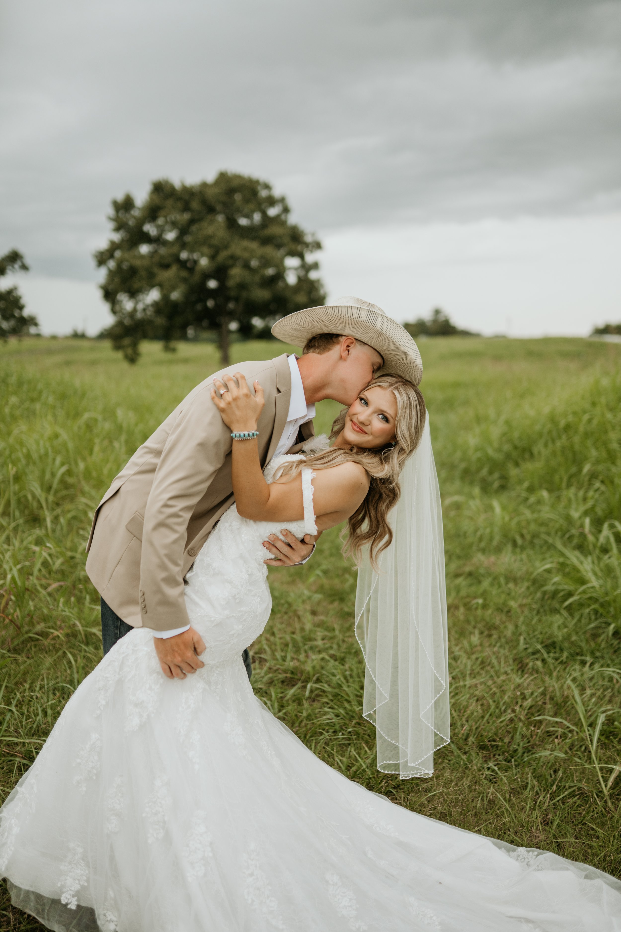 A bride and groom in wedding attire in a grassy field. The groom is wearing a beige suit and cowboy hat, kissing the bride on her forehead. The bride is in a white wedding gown with lace details, smiling at the camera, with long blonde hair and a veil. Overcast sky and trees in the background.