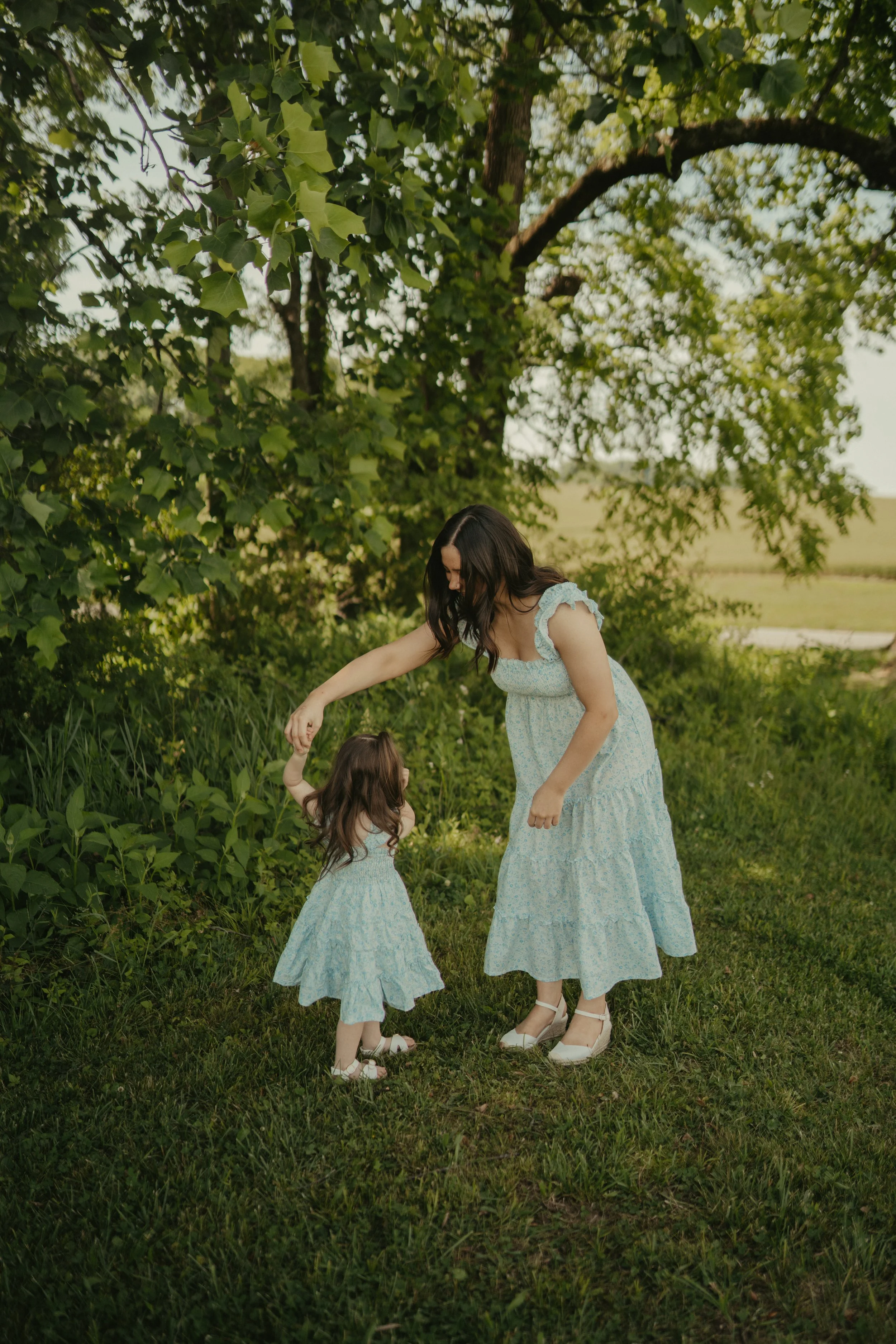 A woman and a young girl in matching light blue floral dresses holding hands in a grassy outdoor area with trees and open fields in the background.
