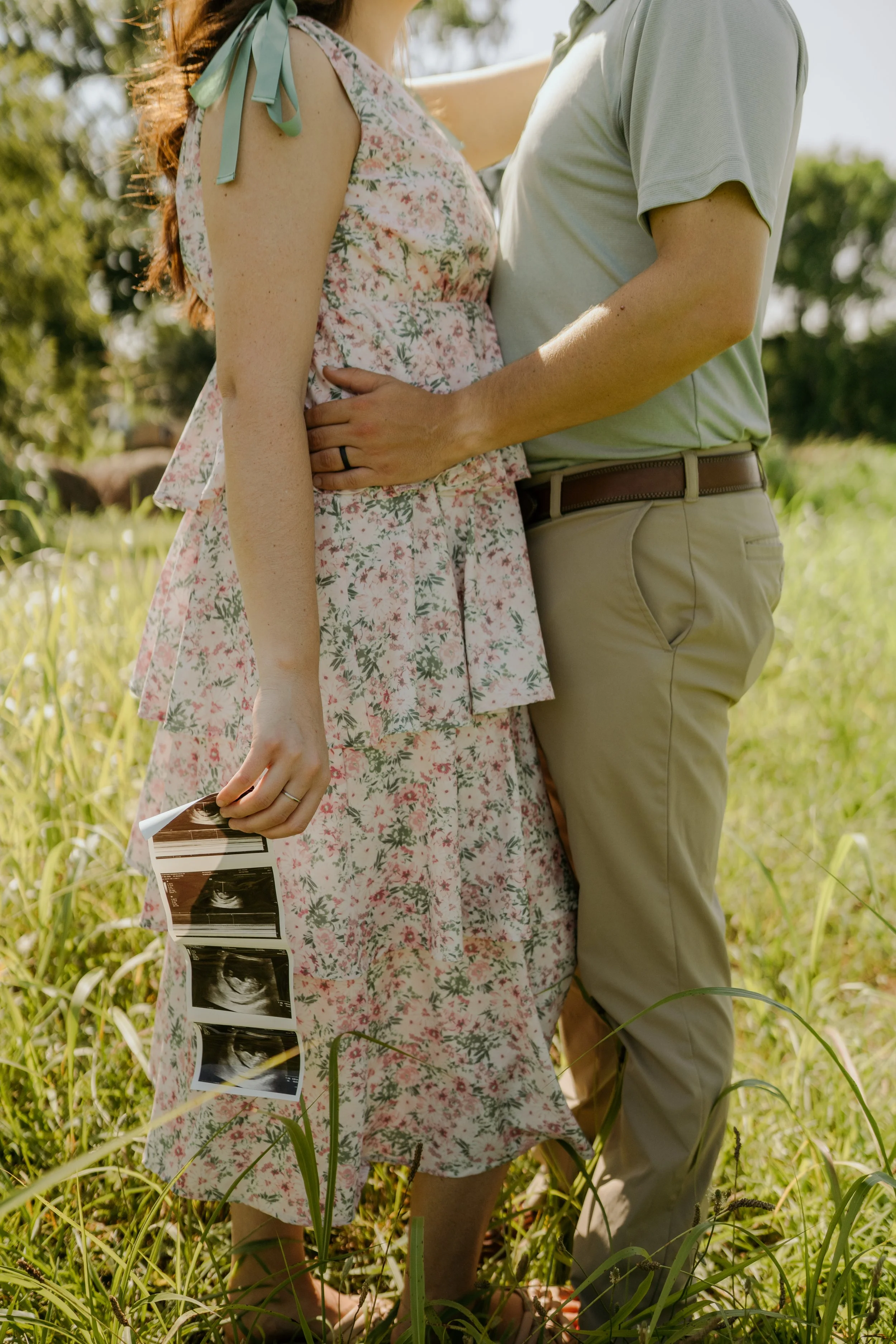 A pregnant woman in a floral dress holding ultrasound pictures stands next to a man in a light-colored shirt and tan pants, with their arms around each other in a grassy outdoor setting.