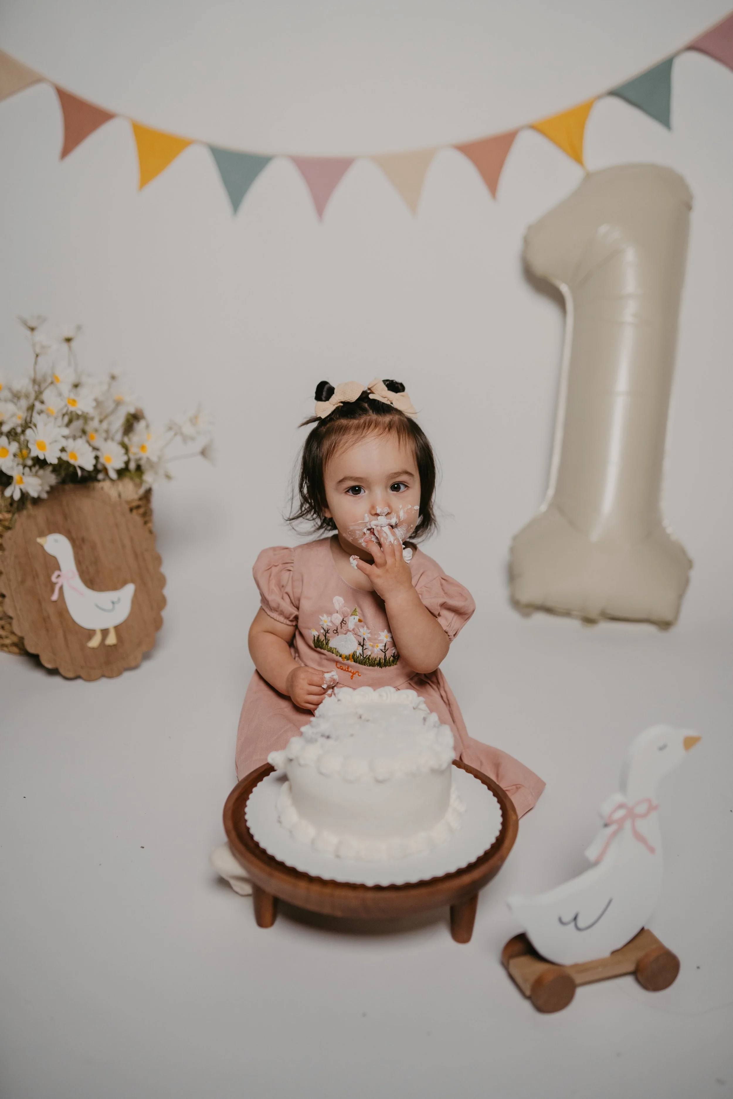 Young girl with dark hair, wearing a pink dress with lamb embroidery, celebrating her first birthday with a cake. She has cream on her face and fingers, and is sitting at a small wooden table with a white cake. Decor includes colorful bunting, a larg