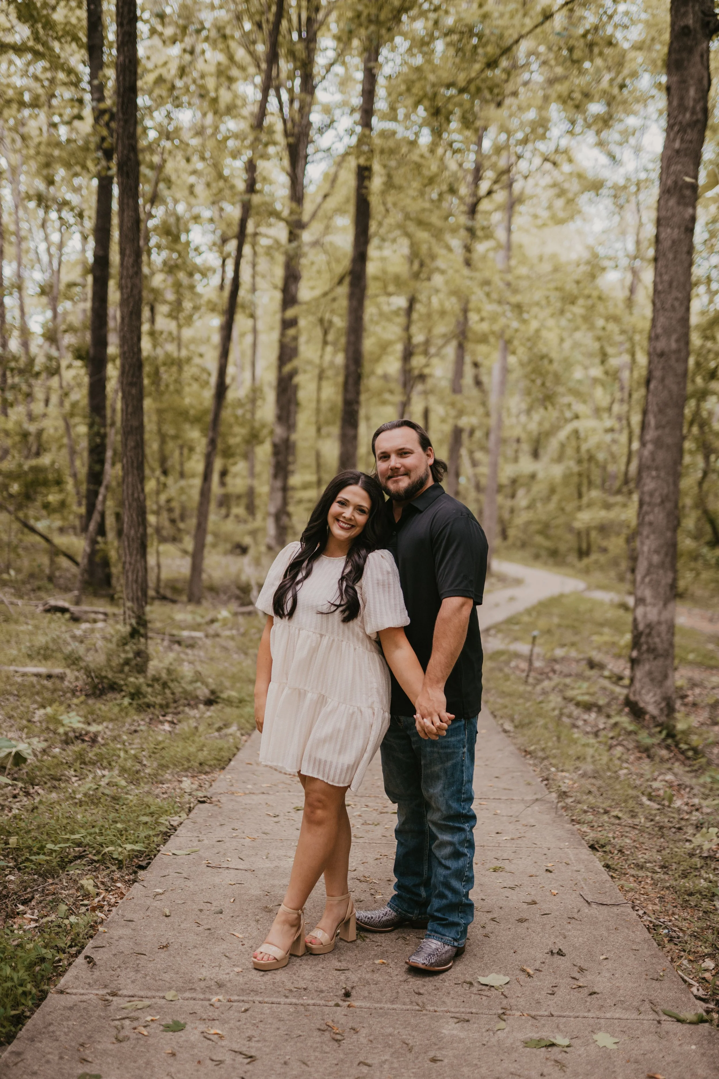 A smiling couple holding hands and standing on a paved walking path in a forested area with tall trees and green foliage in the background.