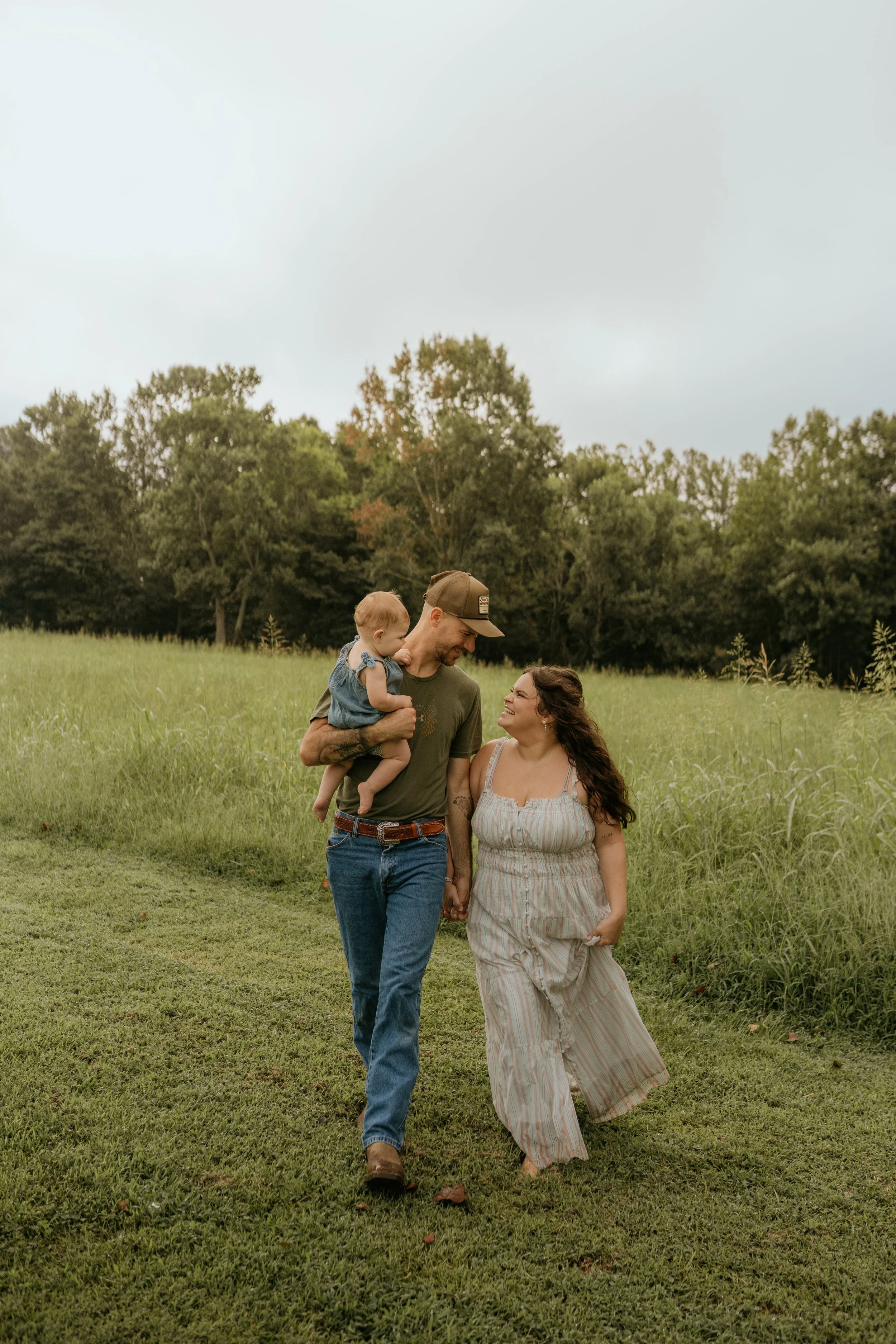 A family of three, a man holding a small child and a woman, walking hand in hand outdoors in a grassy field with trees in the background on an overcast day.