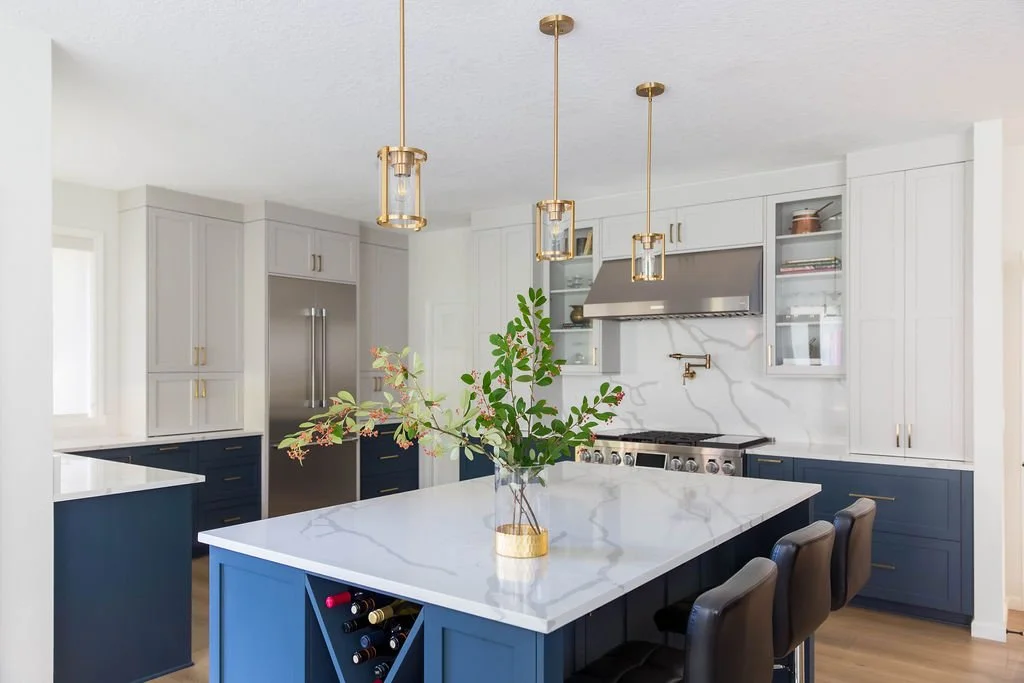 Modern kitchen remodel featuring a navy blue island with waterfall quartz countertop, white shaker cabinets, brass pendant lighting, stainless steel range, and built-in wine storage in a bright, open-concept home.