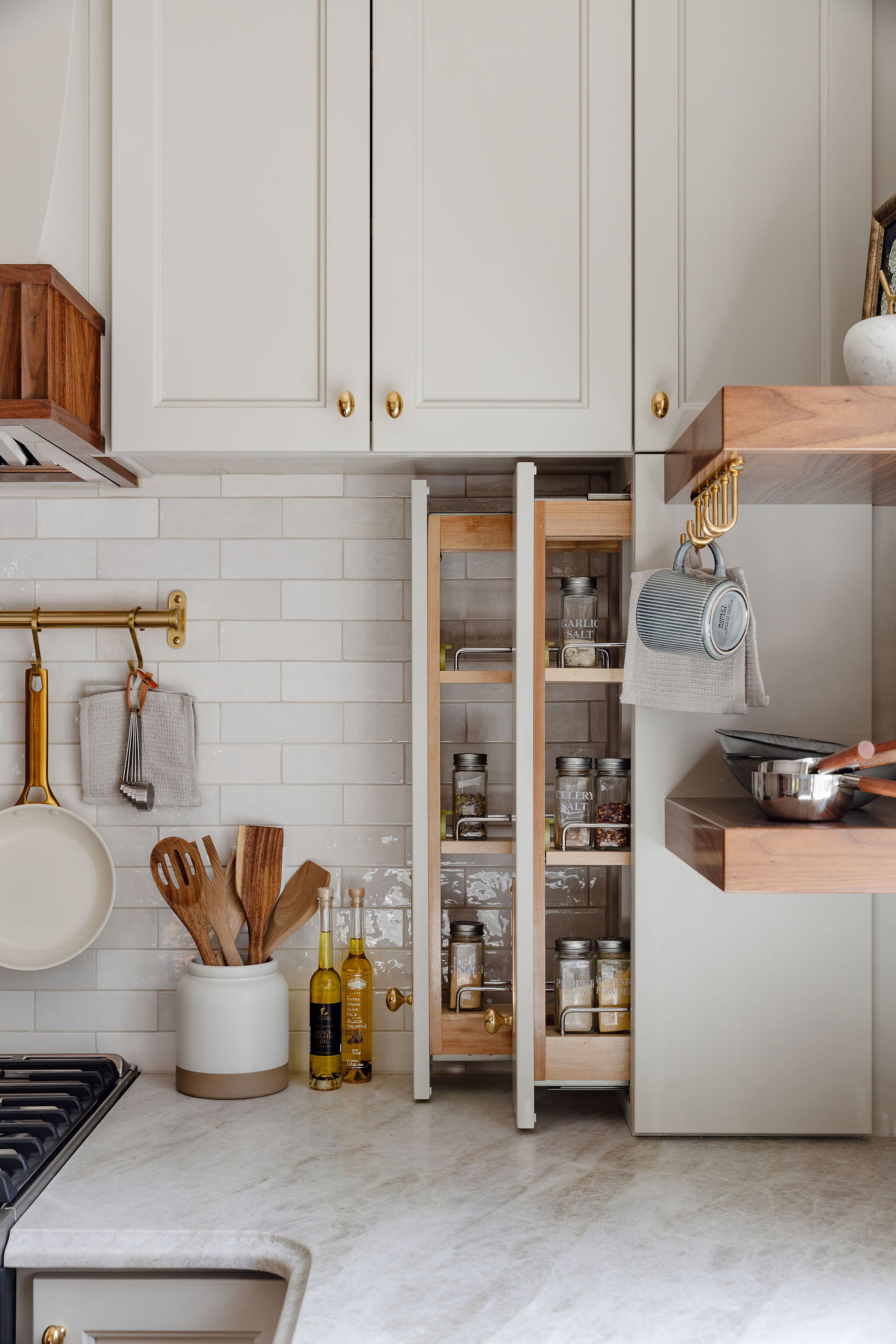 Kitchen Storage spice pull out. Warm white cabinetry with a varrigated white backsplash. Brass cabinetry hardware and warm walnut hood accent and floating shelves. Shelf hooks, Pots and pans hooks. ogee edge Taj Mahal honed countertops. Rejuvenation