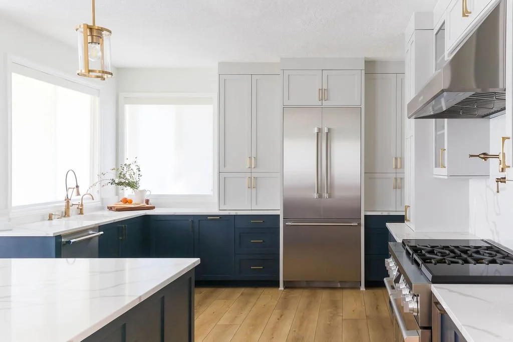 Custom kitchen design featuring a blue island with waterfall quartz countertops, white shaker cabinets, brass pendant lighting, stainless steel range, and open shelving in a bright, modern home.