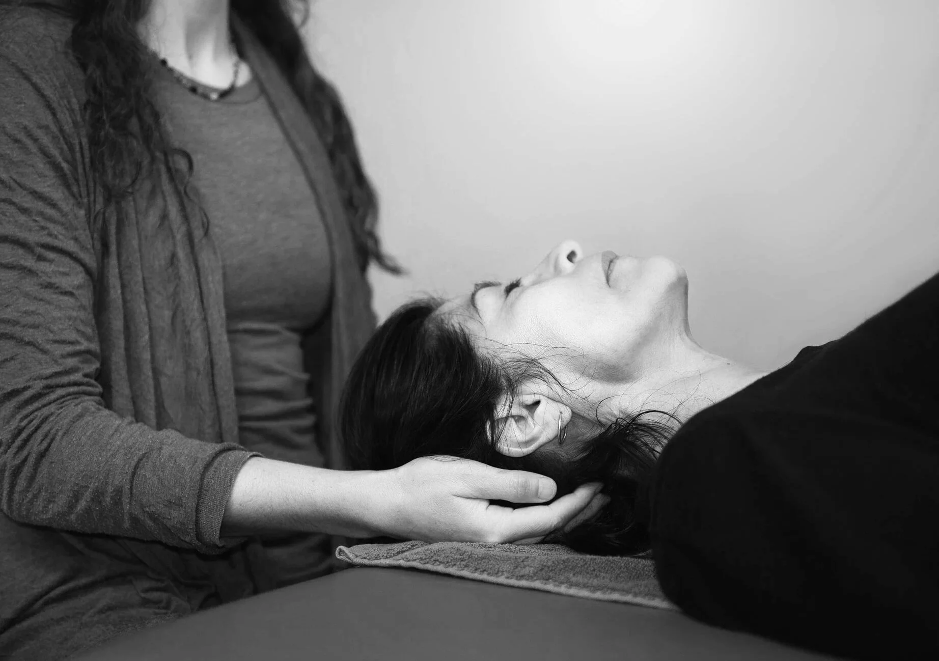 A woman receiving a hands on clinical somatic session from a practitioner lying on a massage like table in black and white.
