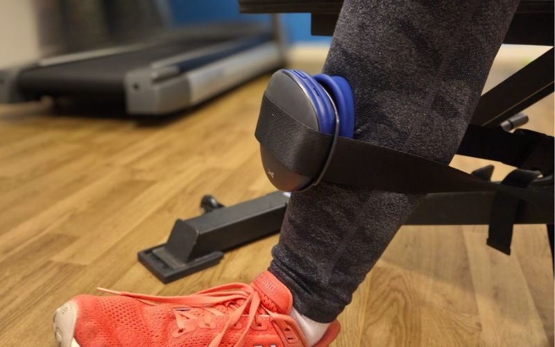 A person's leg with a red athletic shoe resting on a wooden floor, and a blue wireless headphone hanging on the ankle with a black strap.
