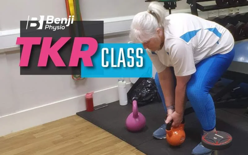 An elderly woman with gray hair tied back, wearing a white T-shirt and blue leggings, lifting a kettlebell in a gym. There are other kettlebells and fitness equipment around her.