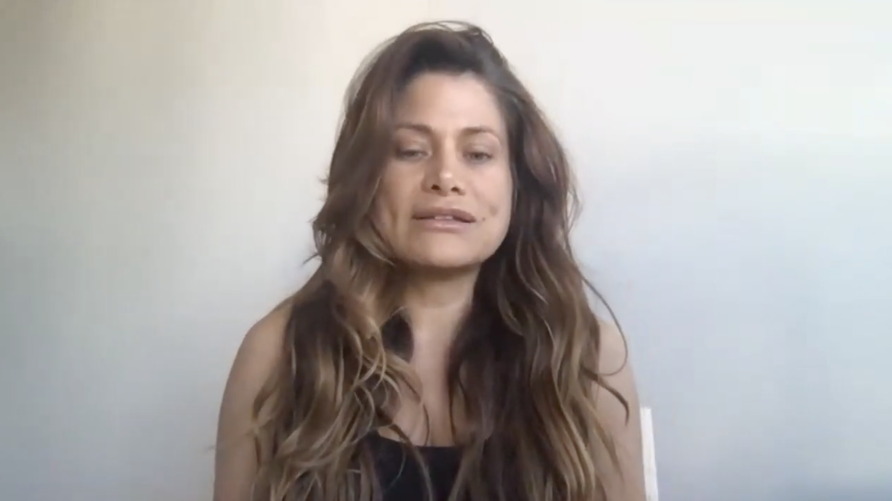 Apollonia Keys with long, wavy brown hair sits against a plain light-colored wall, wearing a black top.