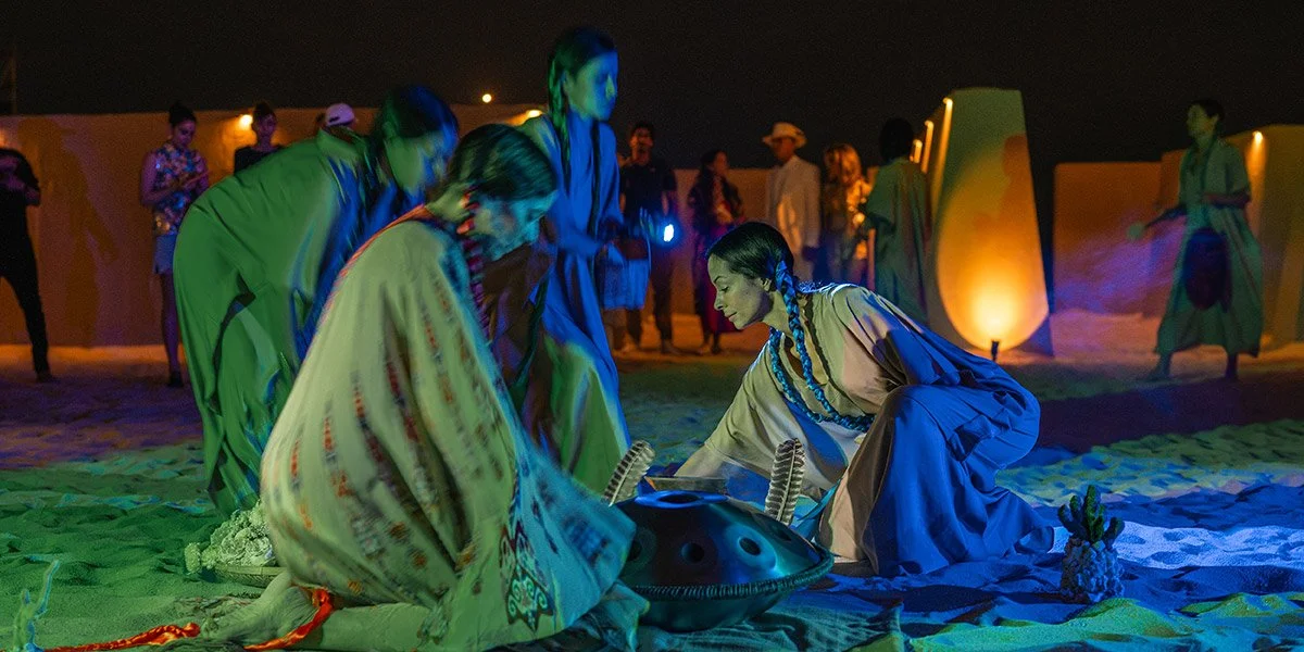 Group of people in traditional Native American attire participating in a nighttime ceremonial dance on a sandy surface, with some standing and others kneeling around a talkative drum, illuminated by colorful stage lights.