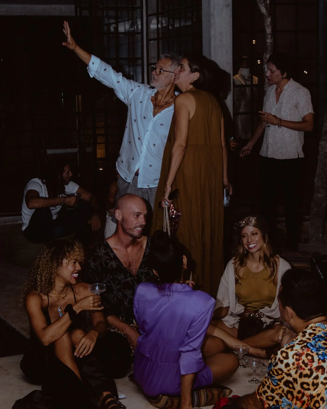 Group of people socializing at an indoor event, some standing and some sitting on the floor, with dim lighting and a relaxed atmosphere.