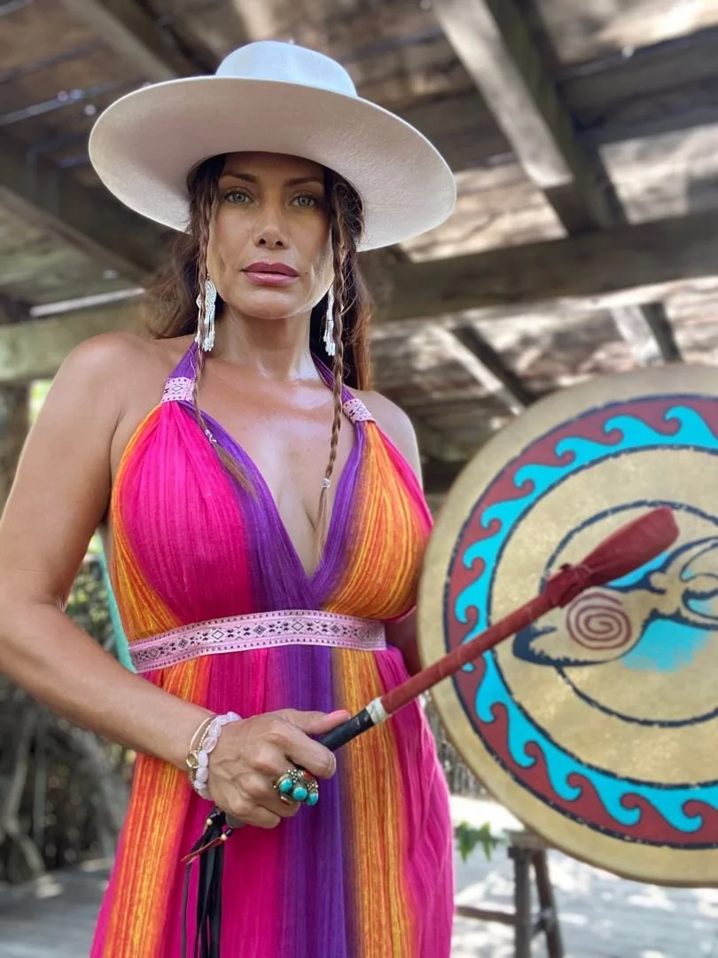 A woman wearing a large white hat and a colorful sundress holding a decorated drum with a drumstick. The background appears to be a rustic outdoor setting.