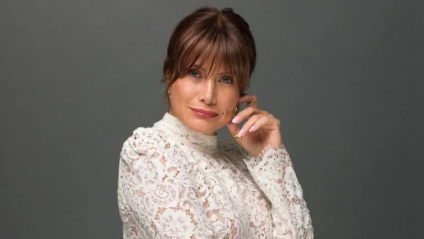 Portrait of a woman with short brown hair wearing a white lace top, posing with her hand near her face against a gray background.