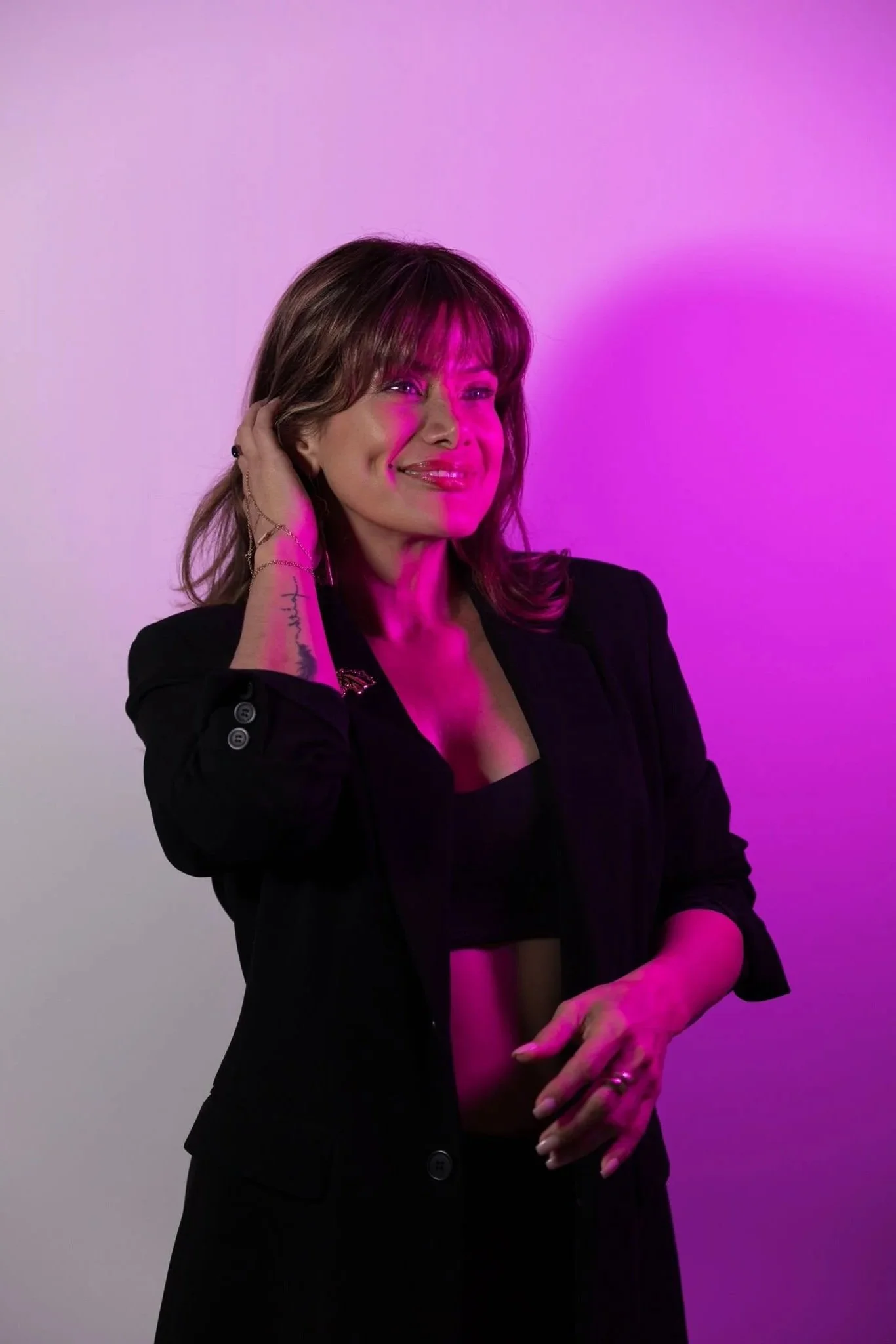 Apollonia Keys with shoulder-length brown hair smiling, wearing black clothing, and illuminated with pink and purple lighting against a plain background.