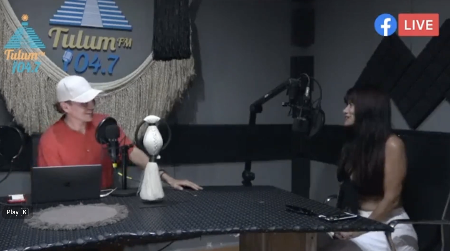 A radio interview in a recording studio with two women, one in a white cap and red shirt, the other Apollonia Keys in a black top. The studio features a wall with the logo of Tulum FM 104.7, microphones, a laptop, and soundproofing panels.