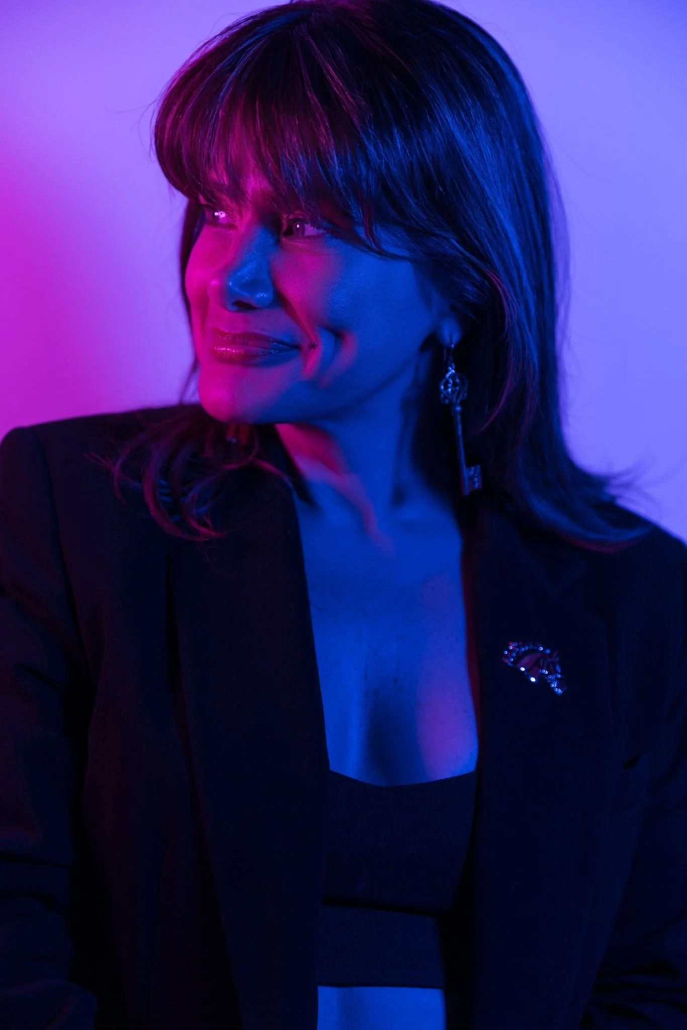 Apollonia Keys with shoulder-length dark hair and bangs is illuminated with vibrant purple and blue lighting, looking to her left with a slight smile, wearing a blazer with a rhinestone pin and long earrings.