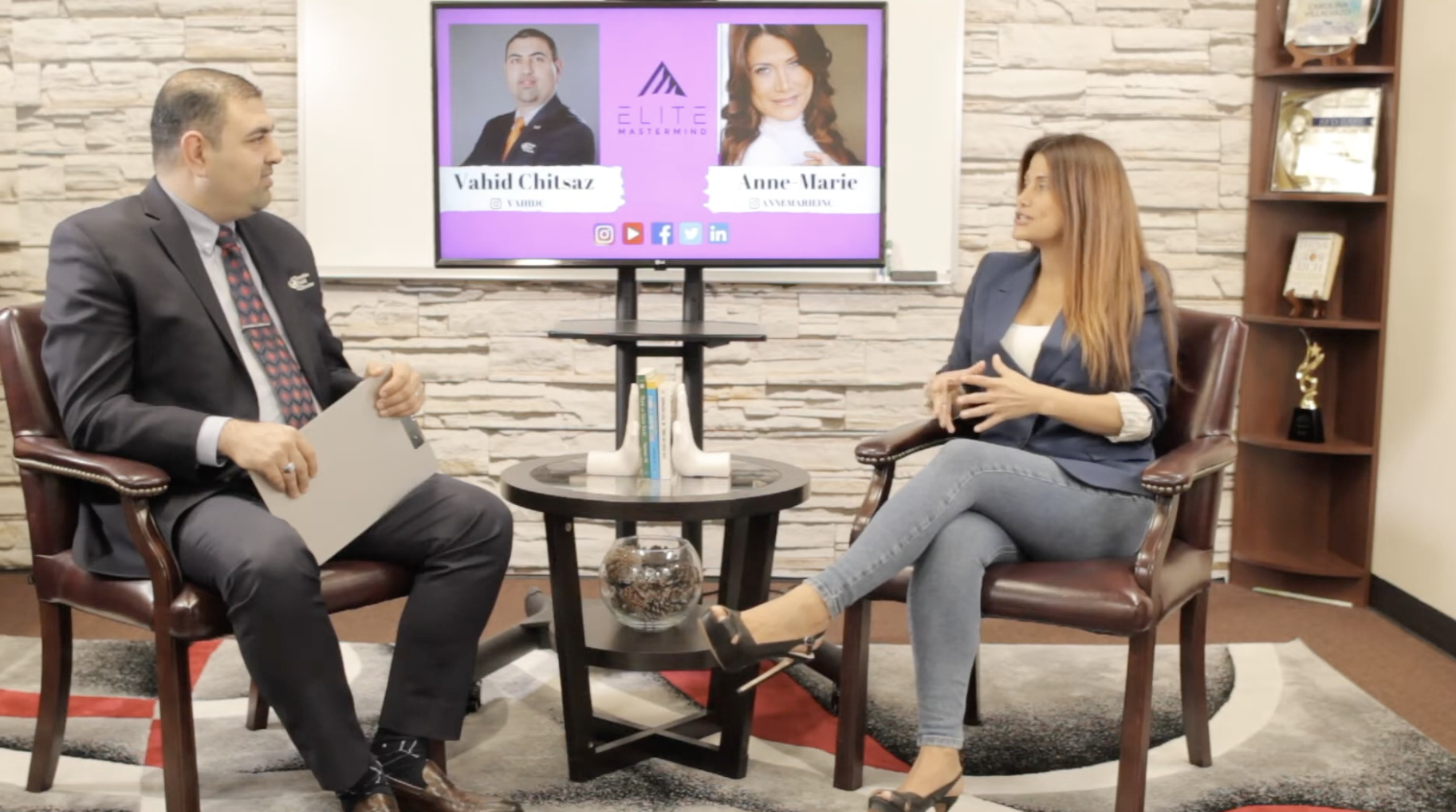 An interview setting with a man in a gray suit and Apollonia Keys in gray jeans and a blue blazer sitting across from each other, with a television screen behind them displaying their names and social media icons.