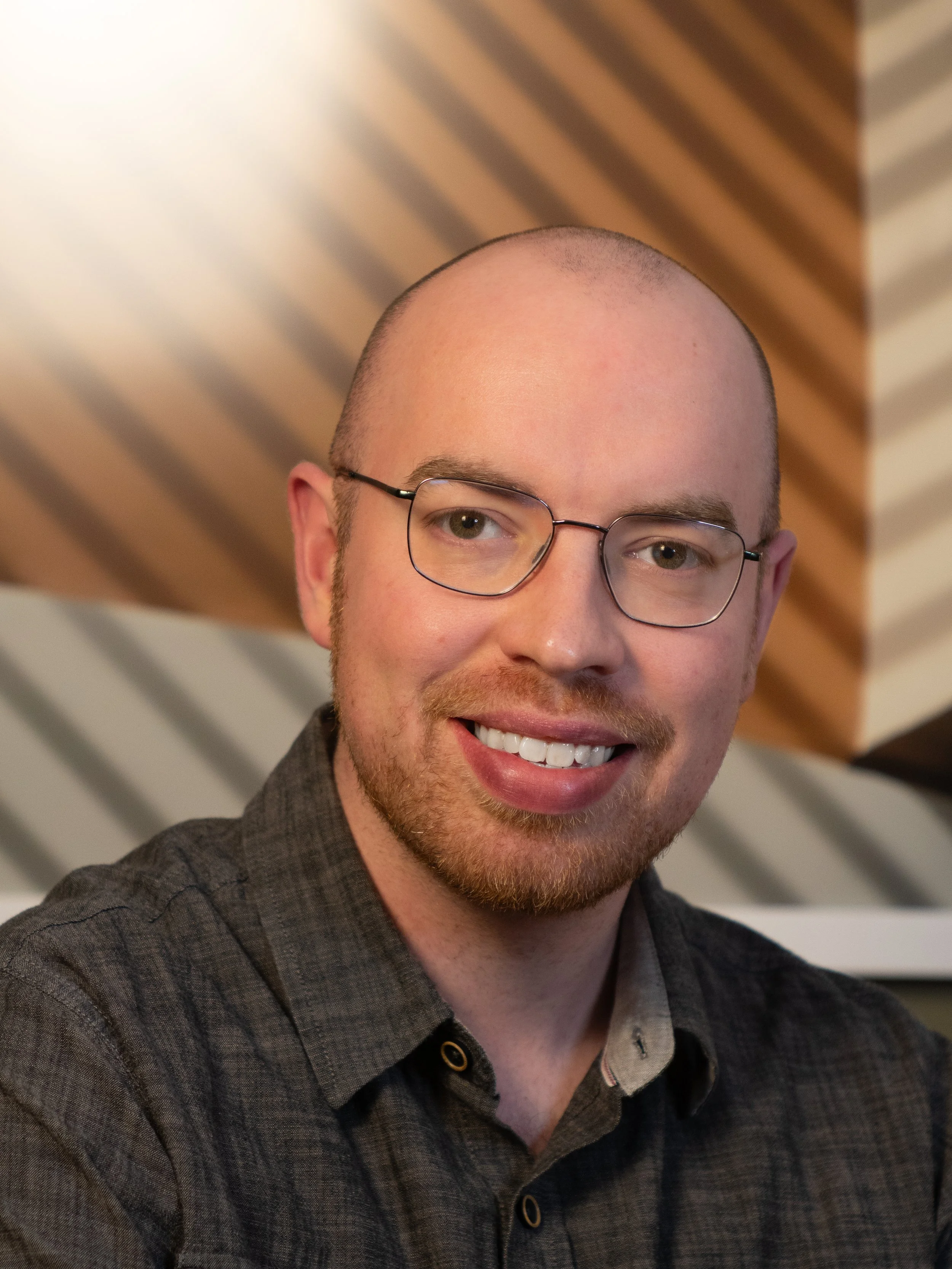 A smiling man with glasses in a gray shirtstanding in front of a brown and beige background.
