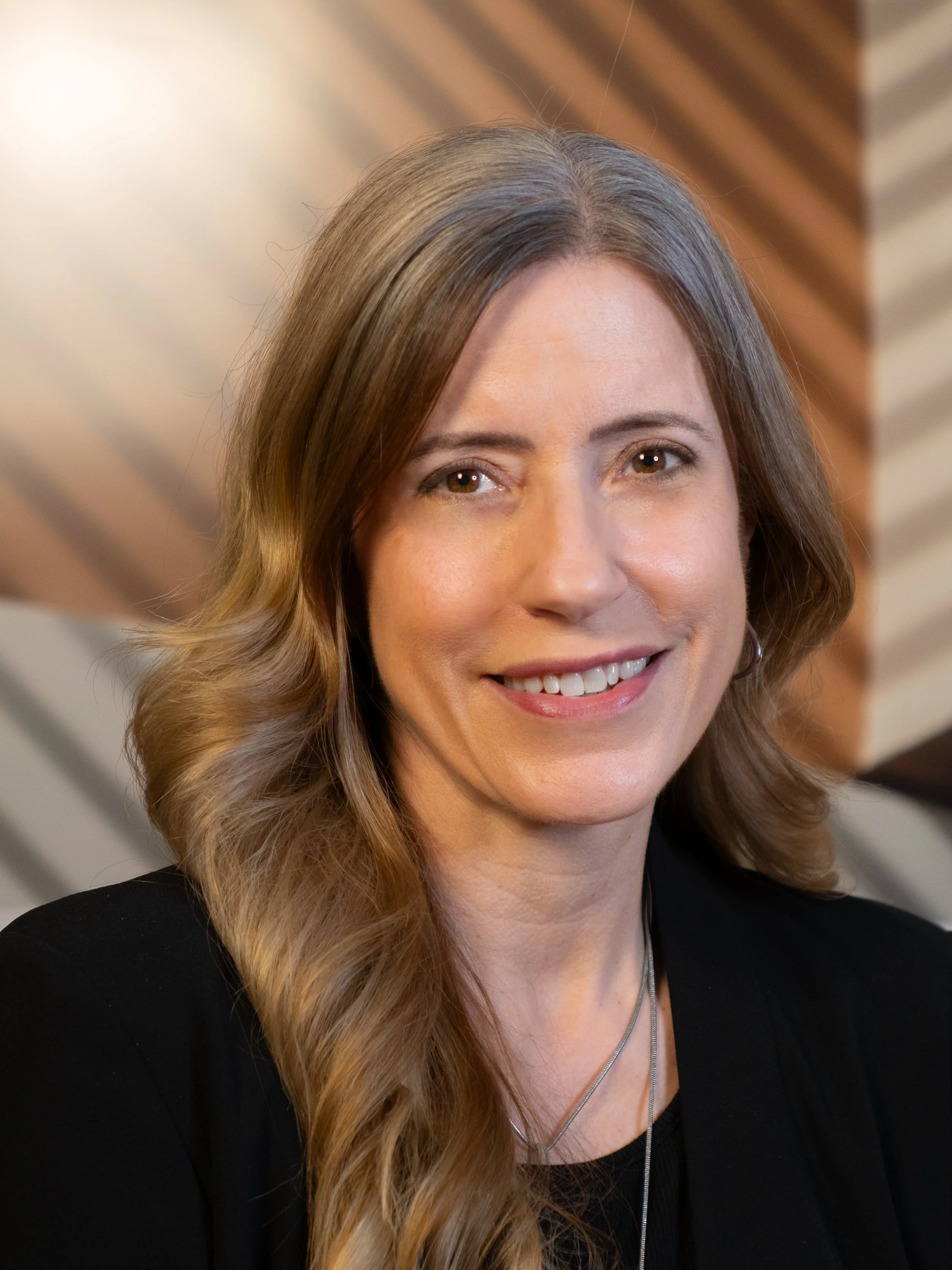 A smiling woman with long blonde hair in a black suit standing in front of a brown and beige background.