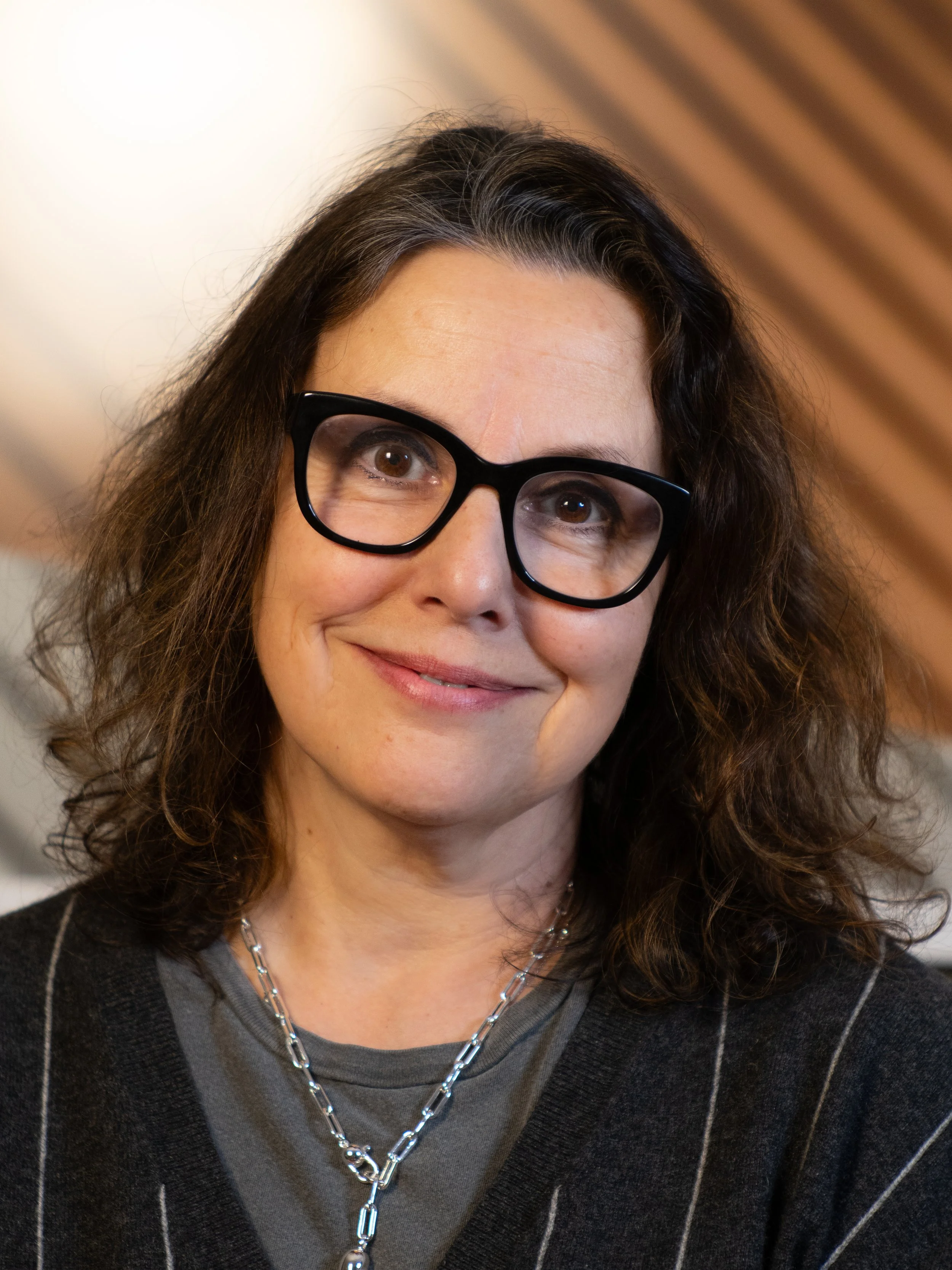 A smiling woman with shoulder-length brown hair and glasses in a gray sweater standing in front of a brown and beige background.