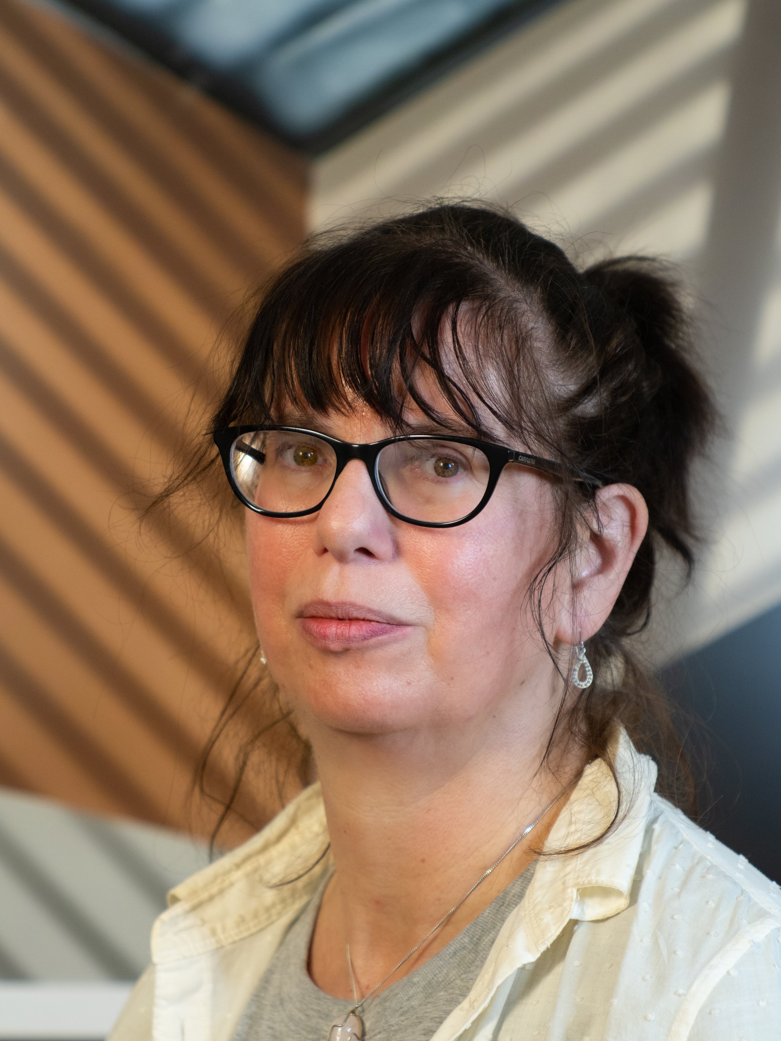 A woman with shoulder-length brown hair wearing glasses and a collared shirt standing in front of a brown and beige background..