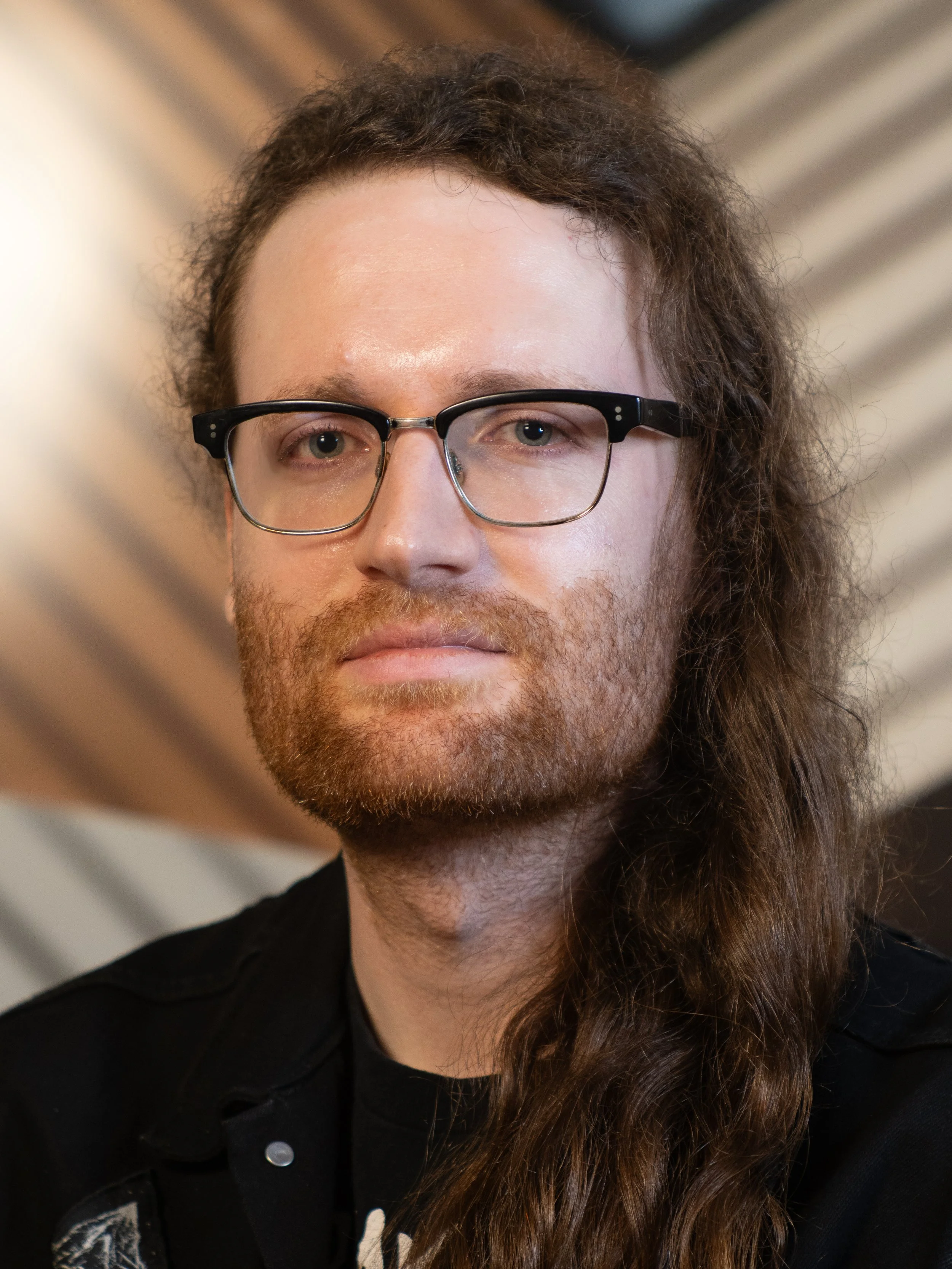 A man with long brown hair and glasses standing in front of a brown and beige background.