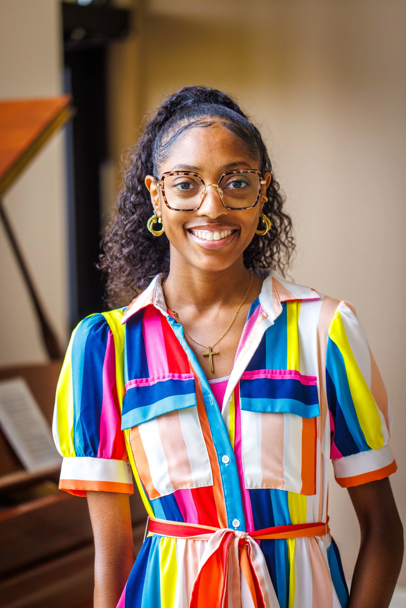 A young woman with glasses and curly hair tied back, smiling, wearing pink earrings, a black blazer, and a black T-shirt with colorful pink and green designs.