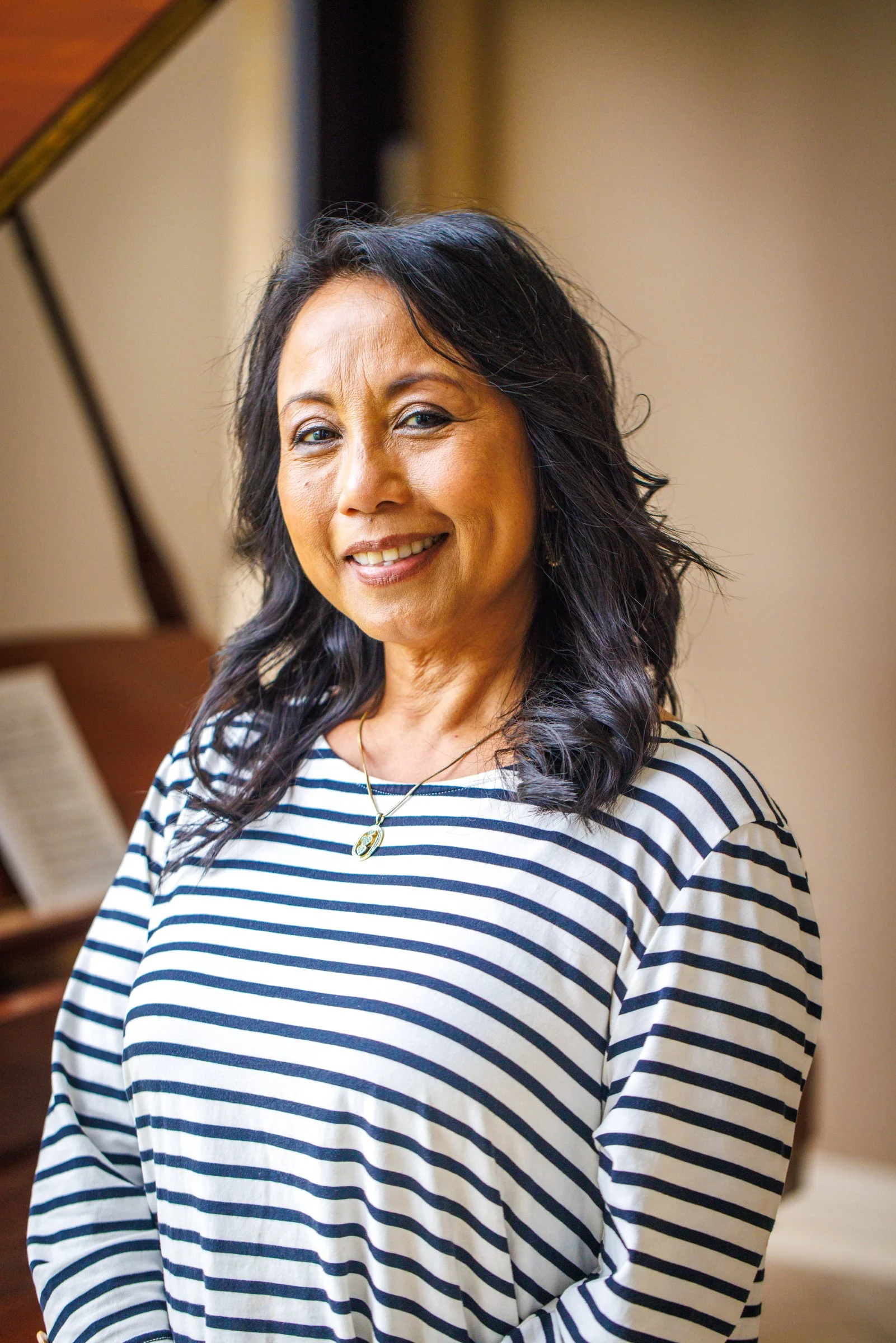 A woman with dark wavy hair smiling, standing indoors in front of a wooden grand piano.