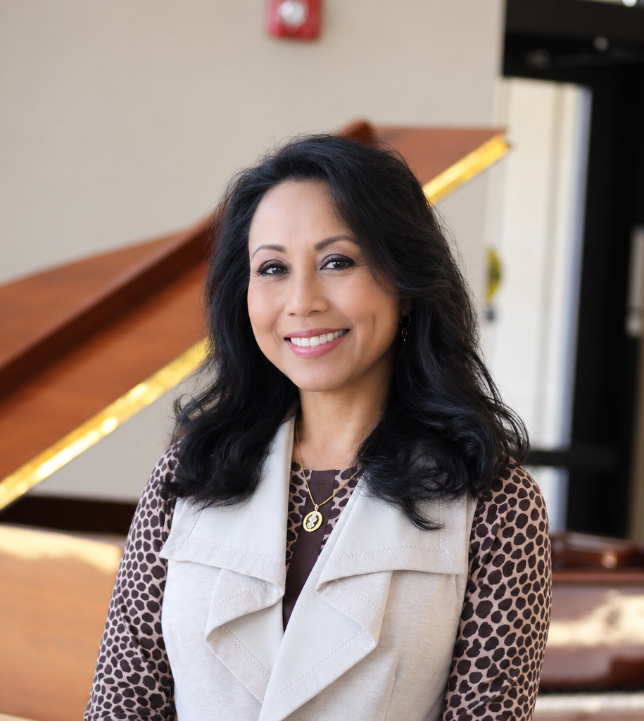 A woman with dark wavy hair smiling, standing indoors in front of a wooden grand piano.
