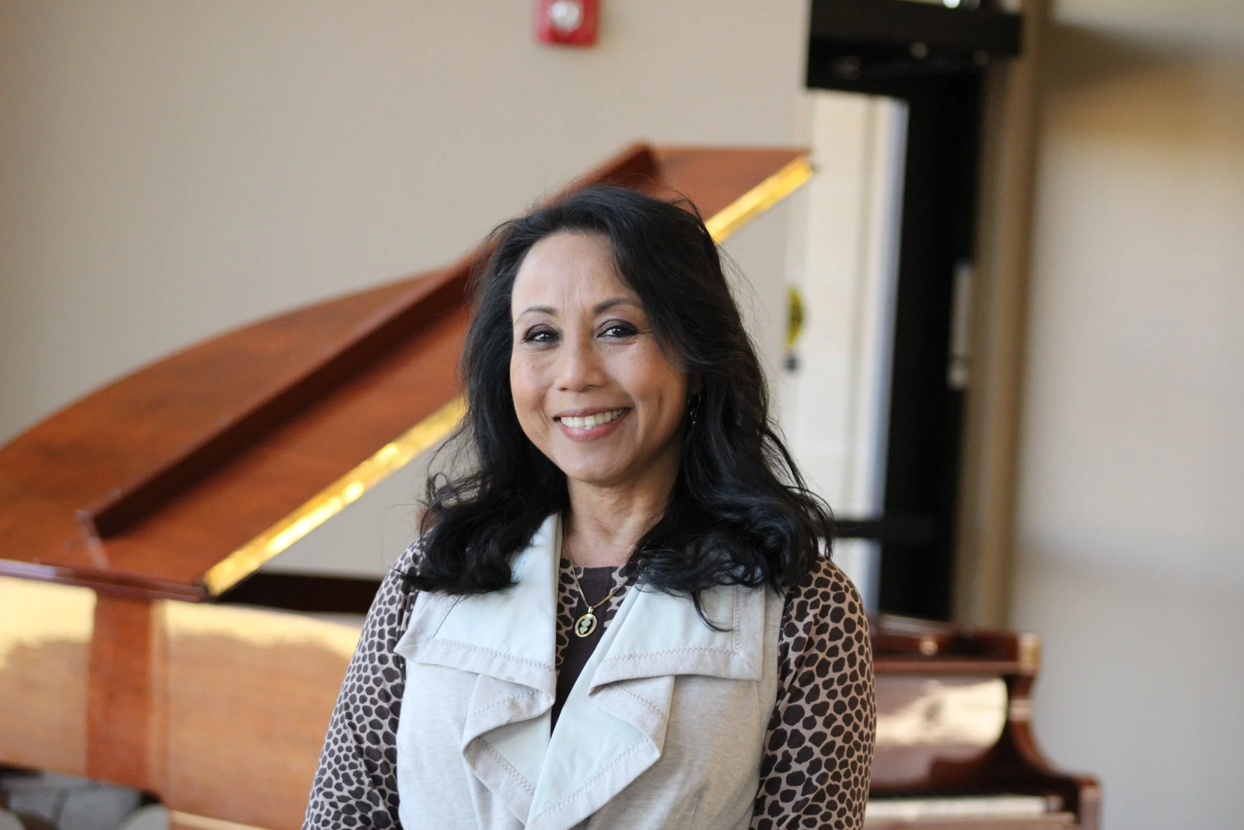 A woman with dark wavy hair smiling, standing indoors in front of a wooden grand piano.