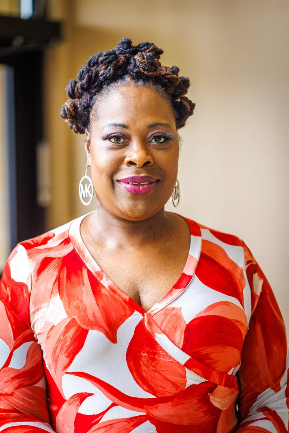 A smiling African American woman with long dreadlocks wearing a purple patterned shirt with hoop earrings.
