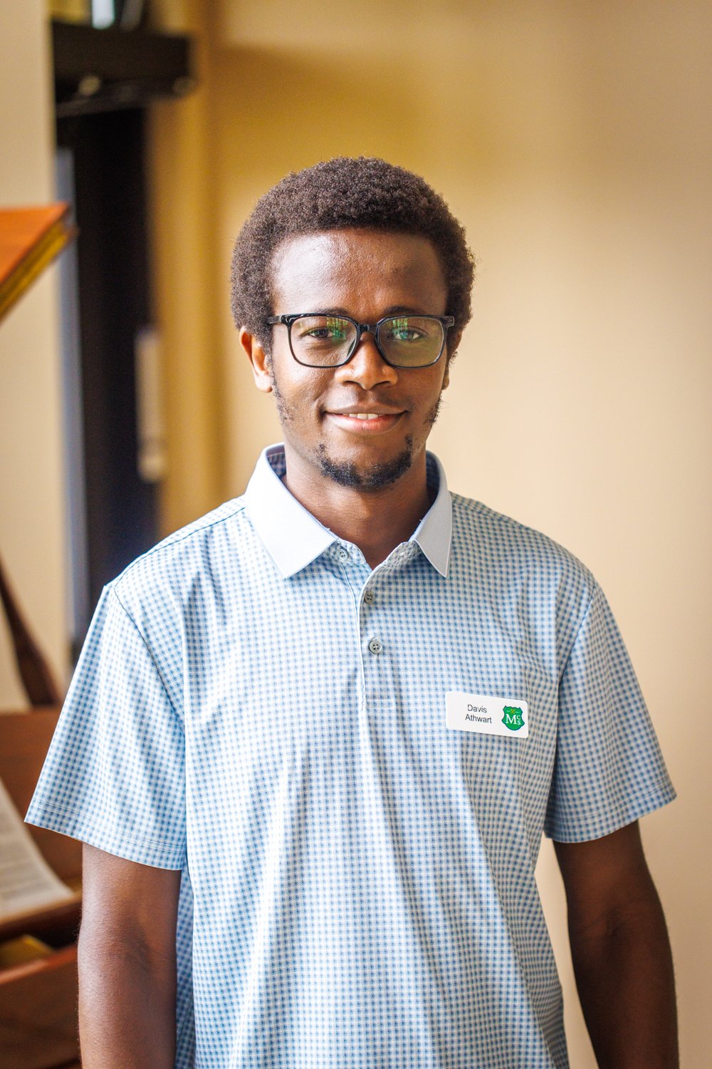 A young man with short curly hair, glasses, and a light blue collared shirt with a name badge that reads 'Davis Athwarm' seen indoors.